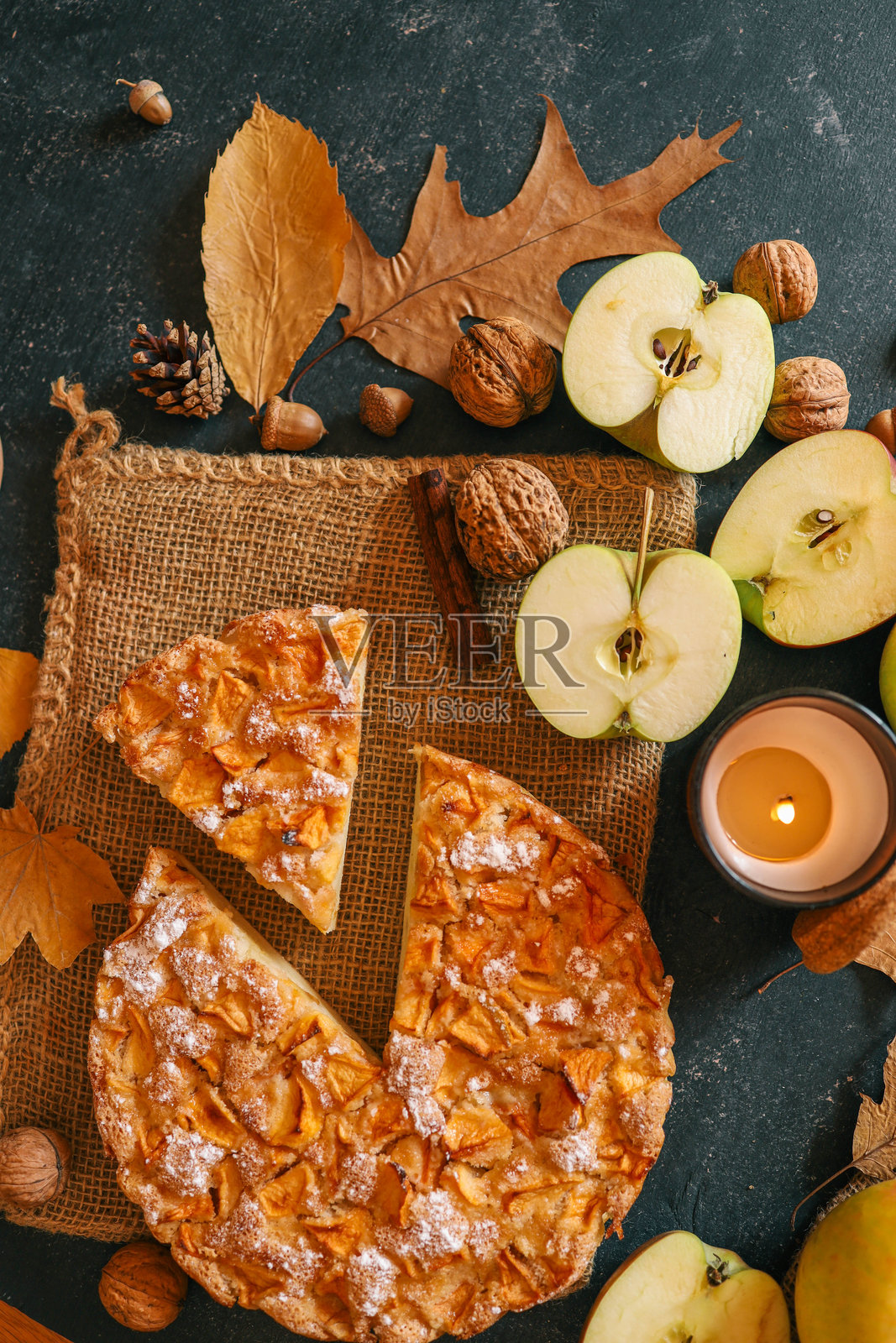 一个温馨的秋季布置，中央是一块新鲜出炉的苹果派，周围摆放着苹果、核桃、一支蜡烛和一杯热饮。照片摄影图片