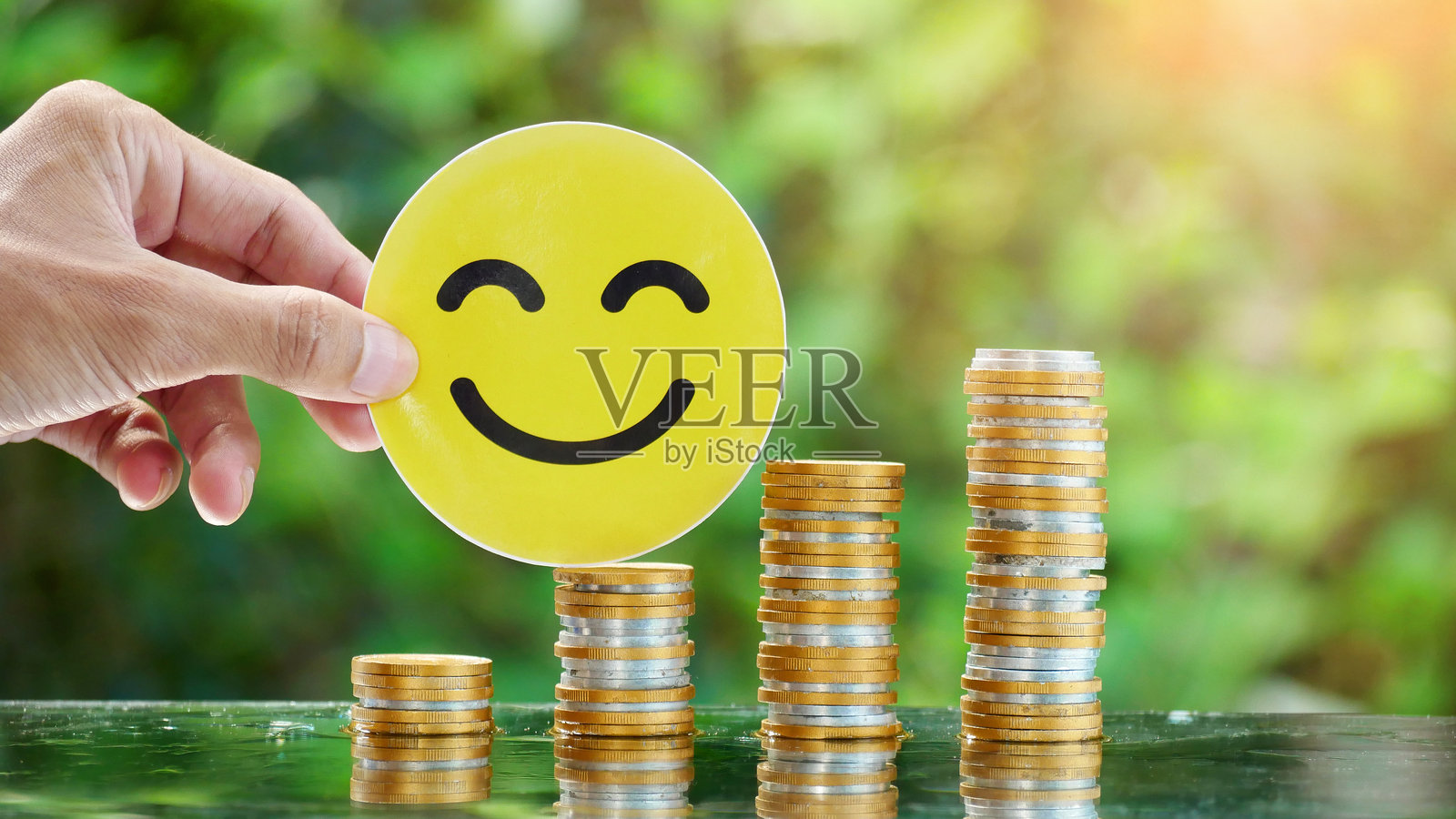 商人手中握着一张微笑面孔的卡片，旁边是一堆硬币。概念照片展示了财务成功、财务自由、经济增长和利润。照片摄影图片