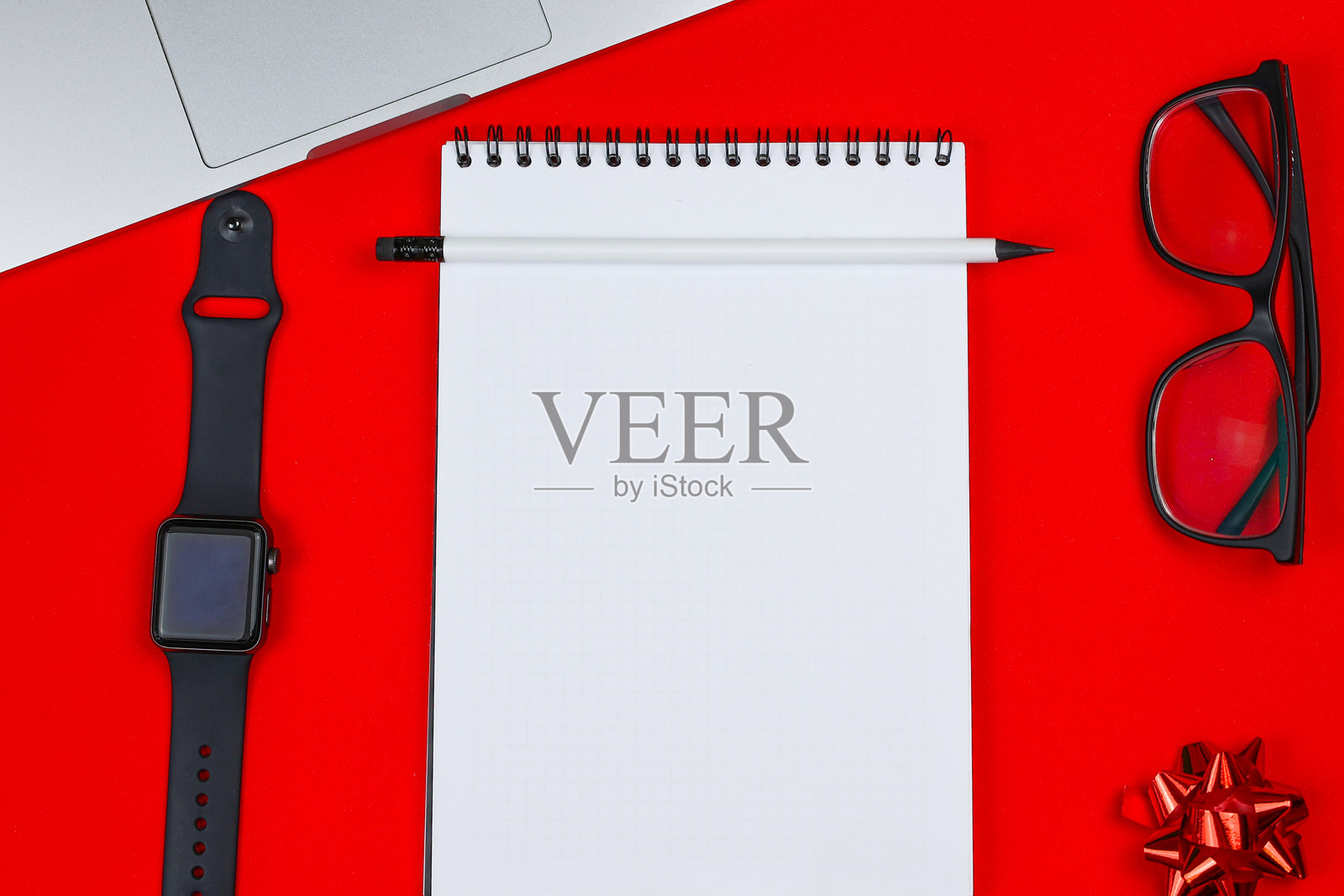 一只白色笔记本配有黑色弹簧，旁边是一支黑色铅芯和橡皮的白色铅笔，置于红色背景的照片中央，周围有一块黑色手腕智能手表、一台笔记本电脑、黑框眼镜和一个节日红色蝴蝶结。照片摄影图片
