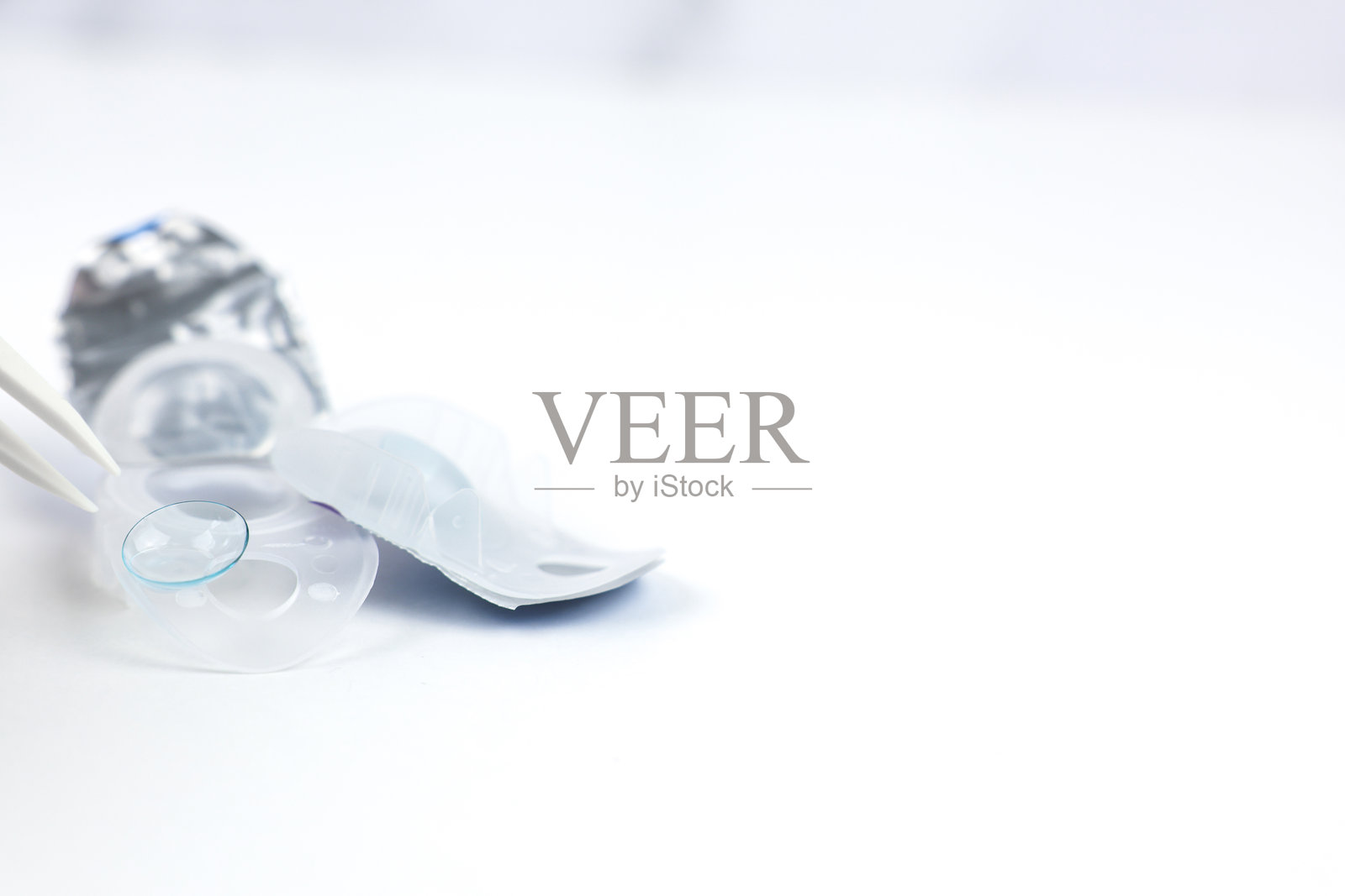 米色镜片夹取镊子伸出，拾起一次性隐形眼镜储存容器边缘的柔软蓝色隐形眼镜。视觉、视力矫正、眼科学的概念。照片摄影图片