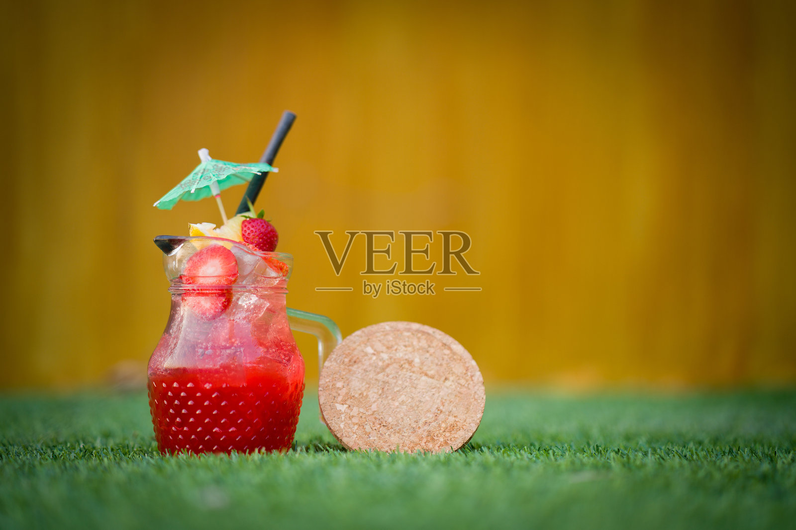 清凉的草莓汁，盛在带有柠檬片、草莓片、小伞装饰和木质杯垫的壶中，放置在一片草地上，背景为黄色。照片摄影图片