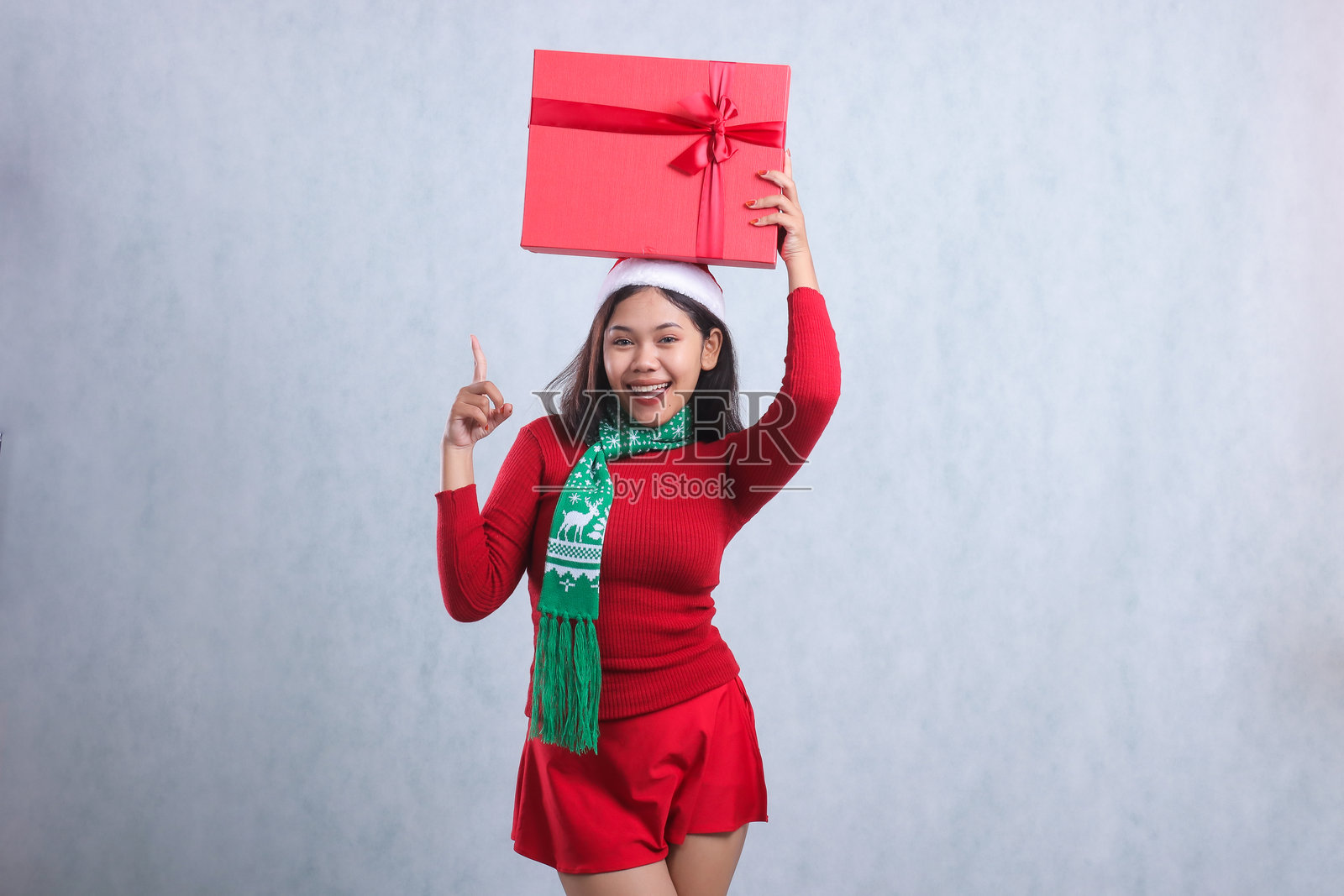 迷人的女性穿着红色圣诞毛衣、圣诞老人帽和围巾，面带愉快的笑容看向镜头，手举着红色的“圣诞快乐”礼物盒并指向上方，背景为白色。照片摄影图片