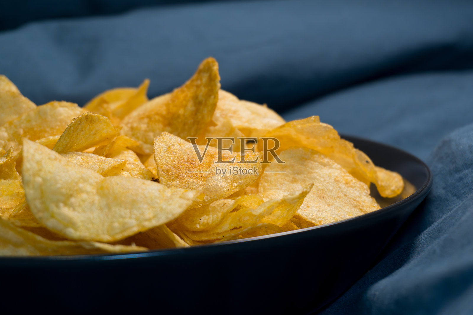 黑碗中的金色薯片：诱人的特写镜头，脆脆的金色薯片盛放在时尚的黑碗中，背景为深蓝色。照片摄影图片