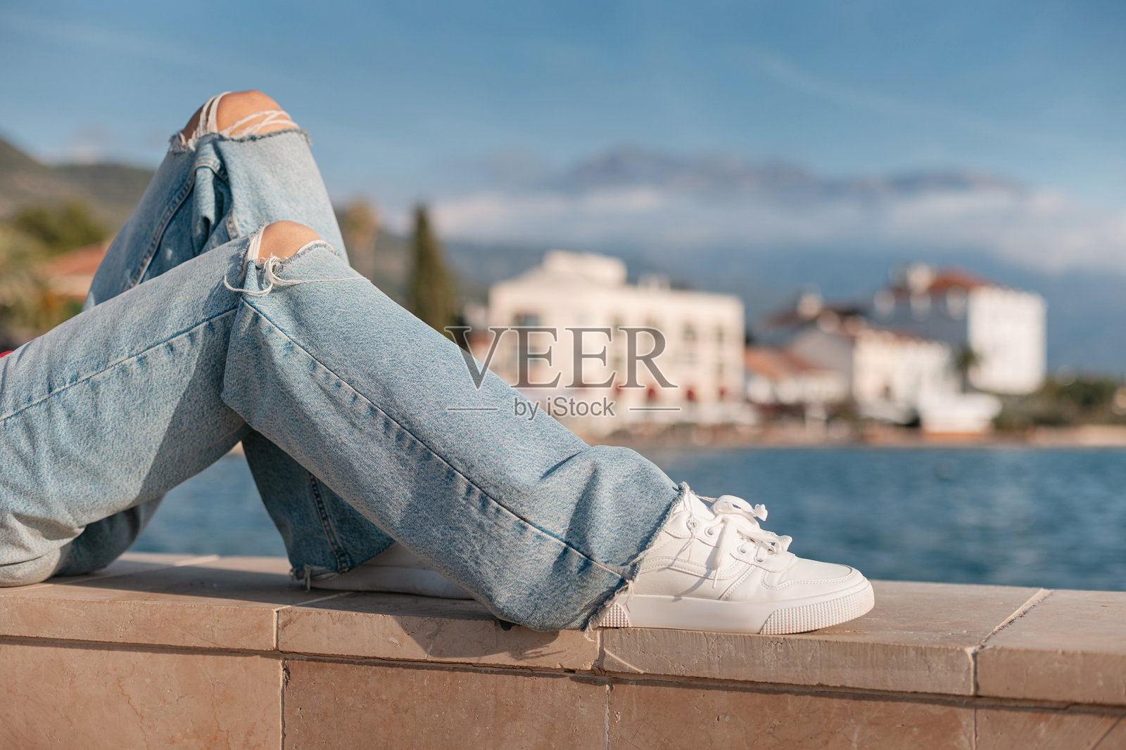 穿着破洞牛仔裤和白色运动鞋的女人坐在海边的矮墙上，欣赏沿海小镇的美景。照片摄影图片