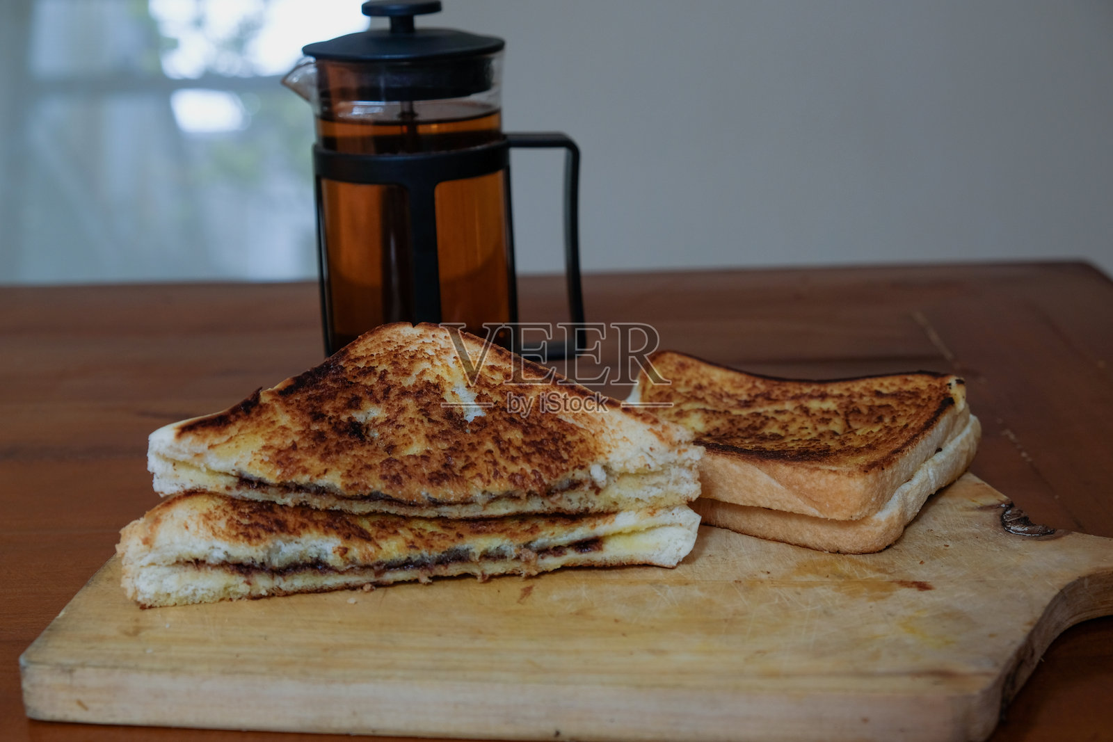 木桌上的切菜板上放着巧克力面包三明治和法式压滤茶壶。照片摄影图片