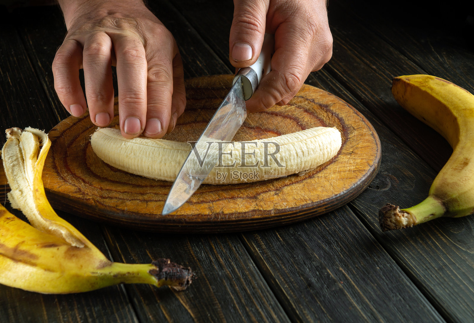 双手熟练地在质朴的木板上切割新鲜的香蕉，周围环绕着成熟的香蕉。温暖的氛围带来一种家的感觉和烹饪创意。照片摄影图片