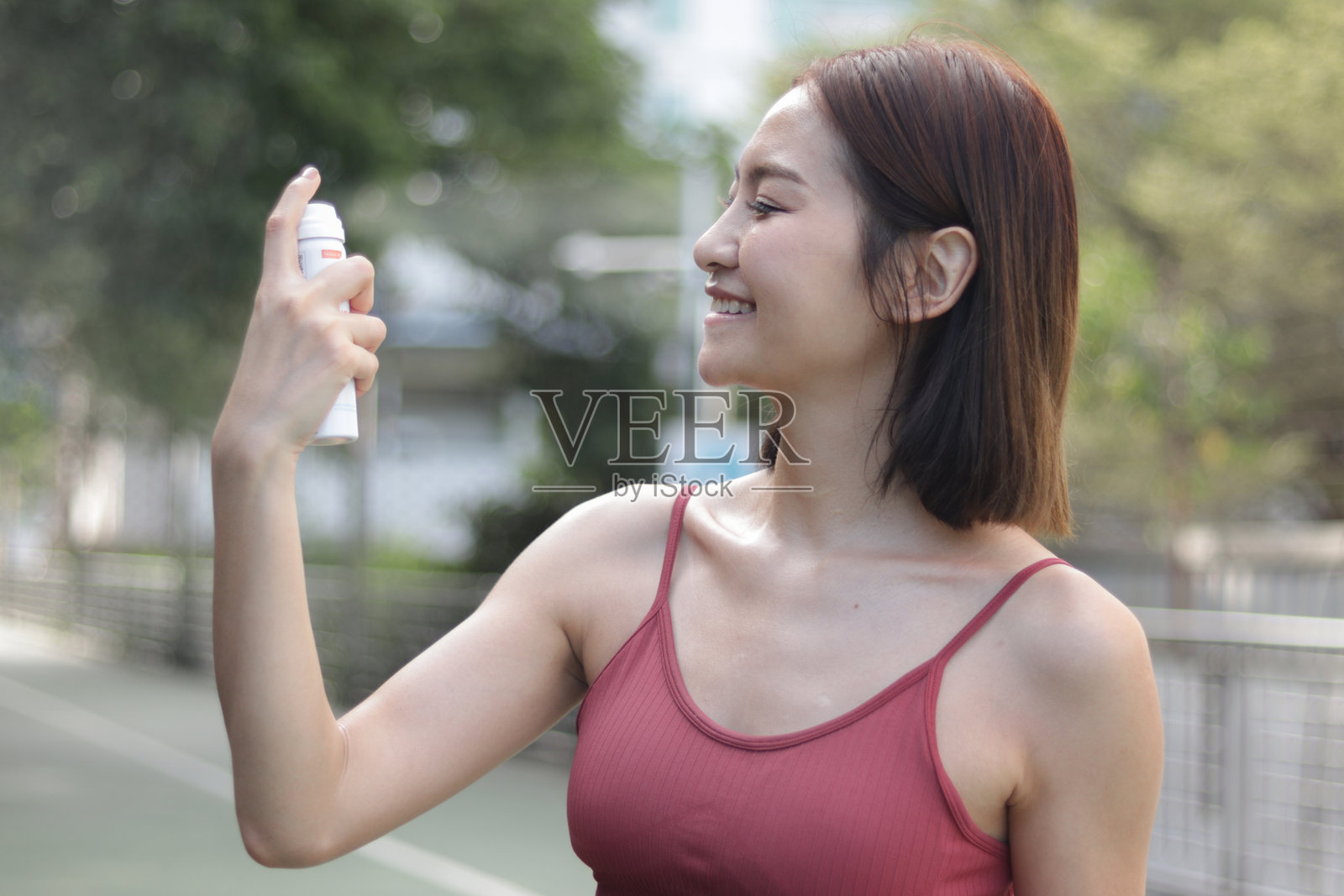 短发运动女孩穿着运动服在阳光明媚的户外慢跑时给脸部喷涂防晒喷雾。亚洲女性在户外锻炼前用防晒霜保护肌肤。照片摄影图片