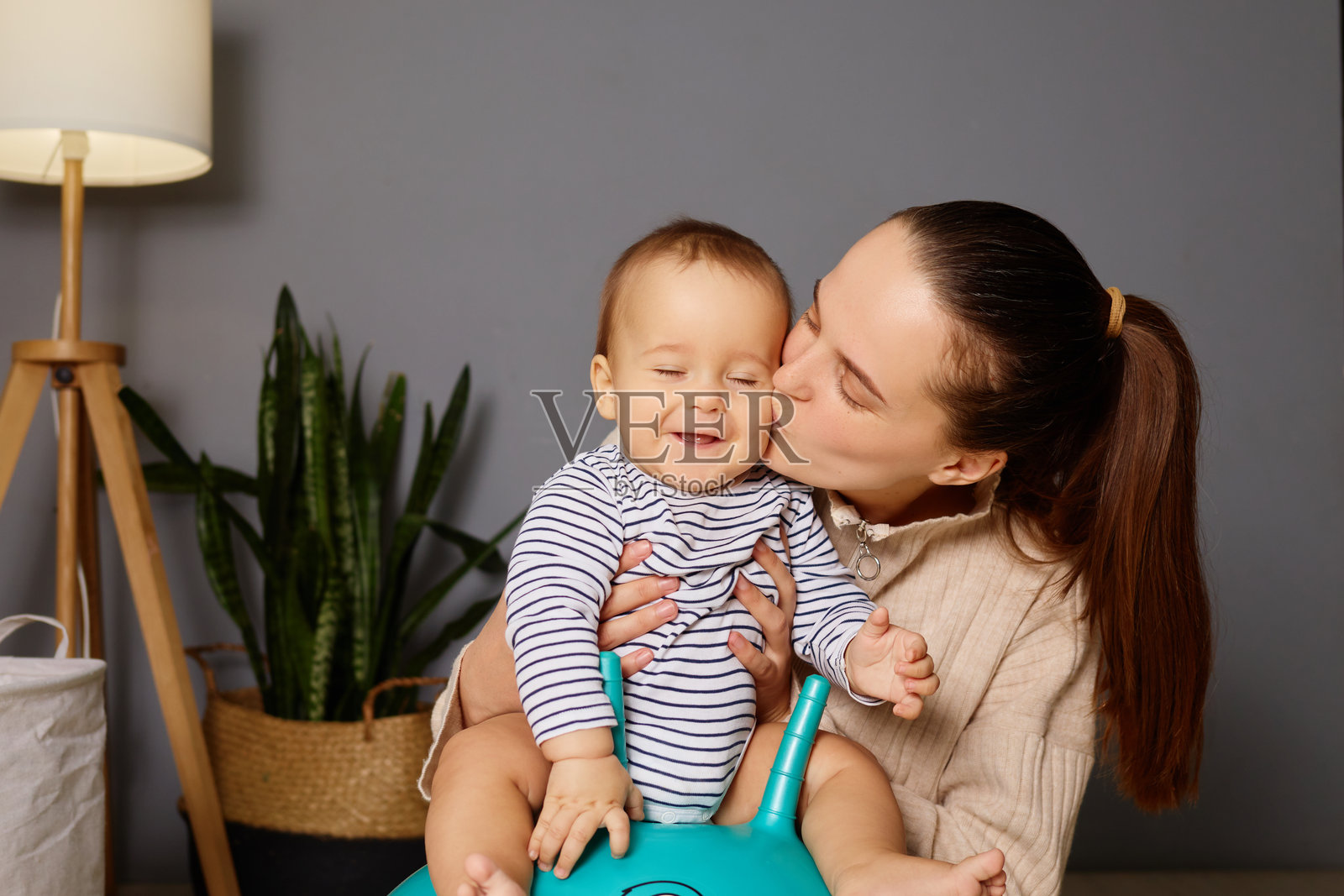 一位母亲在温馨的家中，坐在五彩斑斓的玩具上，温柔地亲吻她快乐的宝宝。植物和柔和的灯光为他们的游戏时光营造了温暖的氛围。照片摄影图片