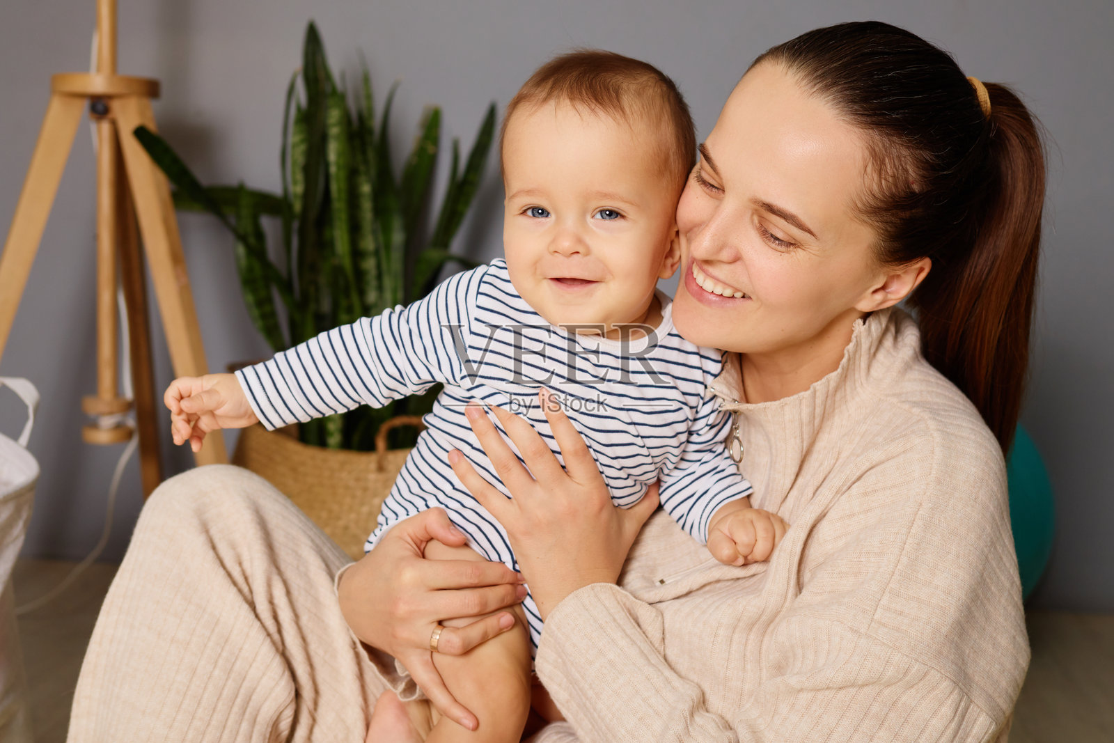一位微笑的母亲在充满绿意的舒适居住空间中抱着她快乐的宝宝。他们分享着愉快的互动，展现出亲密的关系。照片摄影图片