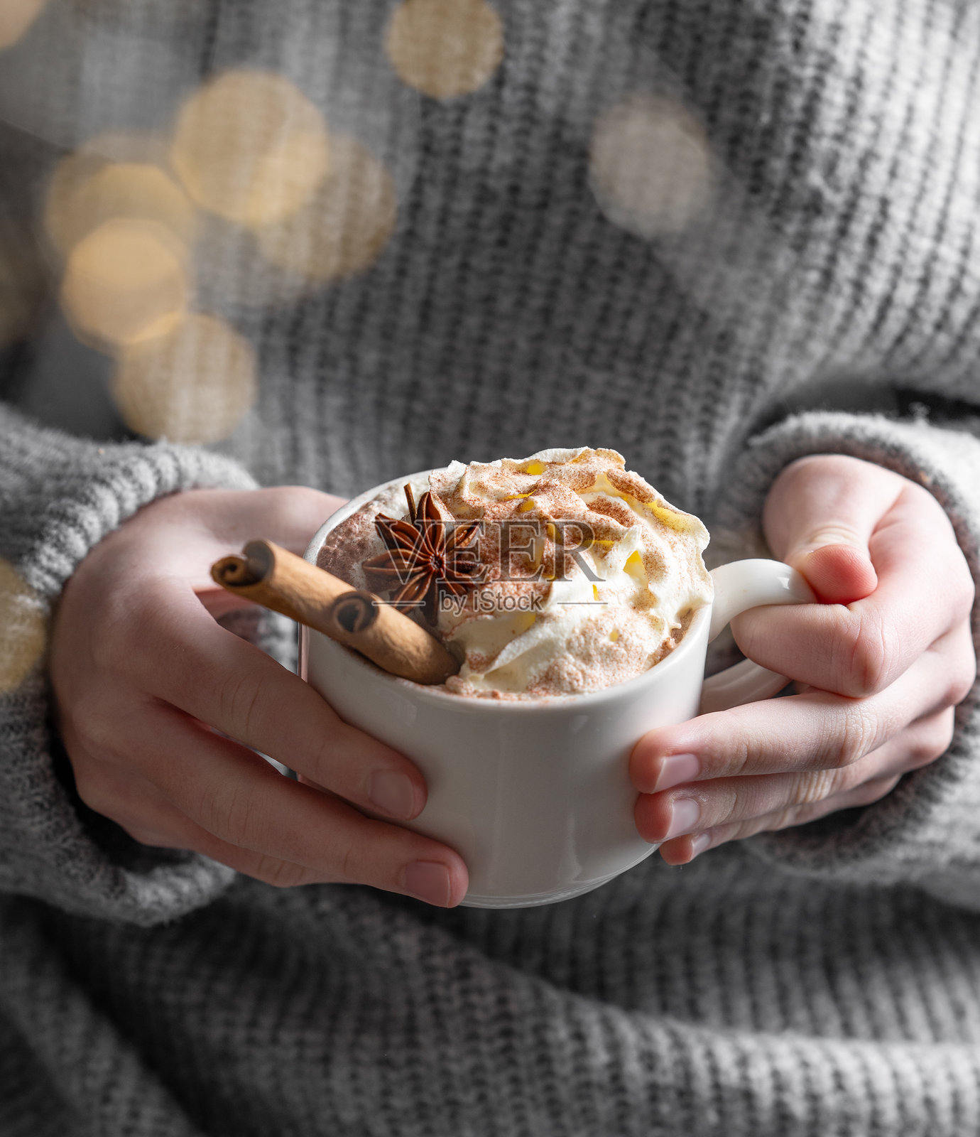 手握热巧克力杯，上面装饰着鲜奶油、肉桂和八角，背景是一件灰色温暖的毛衣。照片摄影图片