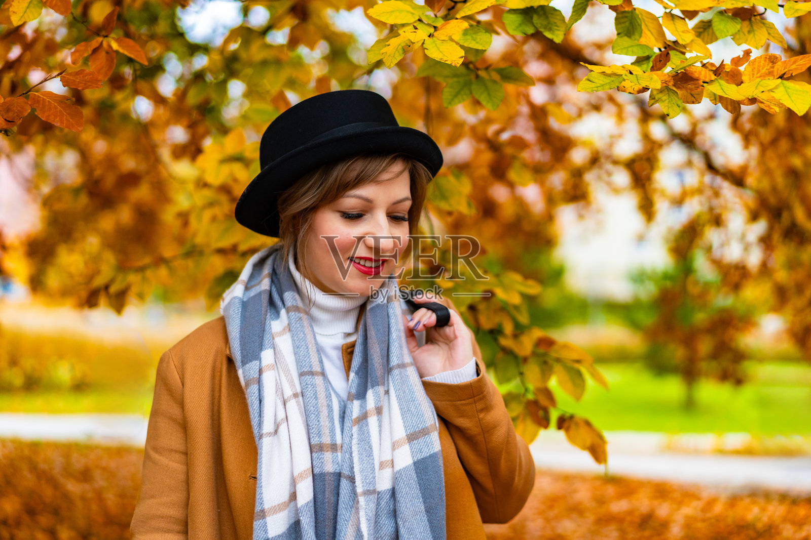 美丽年轻女性的肖像，穿着米色外套，格子温暖围巾，奶油色高领毛衣，黑色边帽，坐在秋天的城市公园里。照片摄影图片