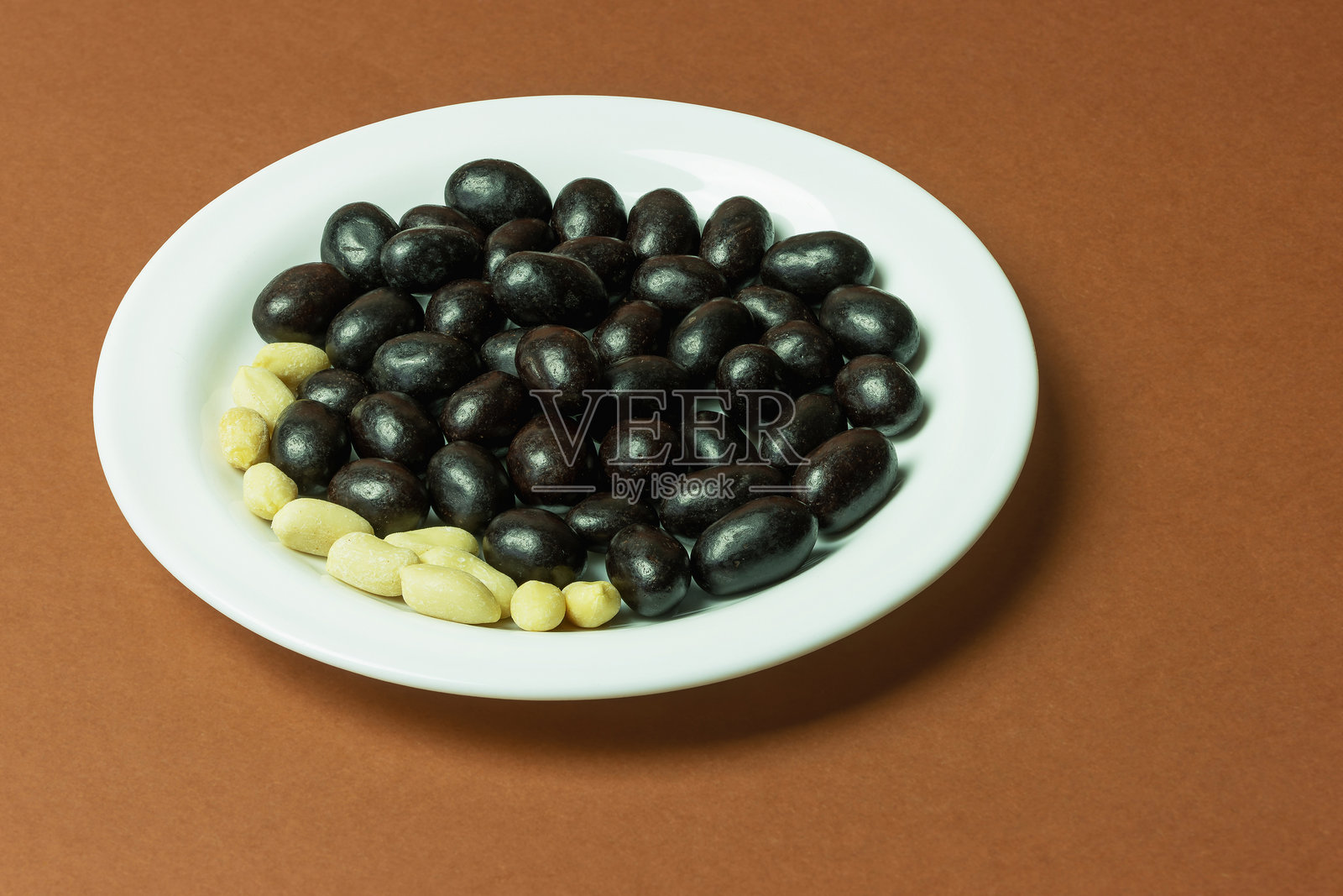 美味的巧克力裹花生整齐地摆放在白色盘子上，背景为棕色。照片摄影图片