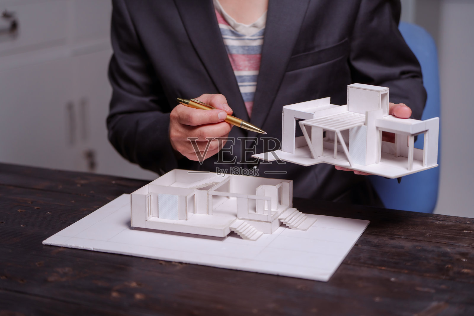一位商人在夜晚的桌子旁坐着，手中拿着一座两层的白色房屋模型。他正在审查设计，草拟计划，并检查屋顶细节，展示现代建筑风格。照片摄影图片
