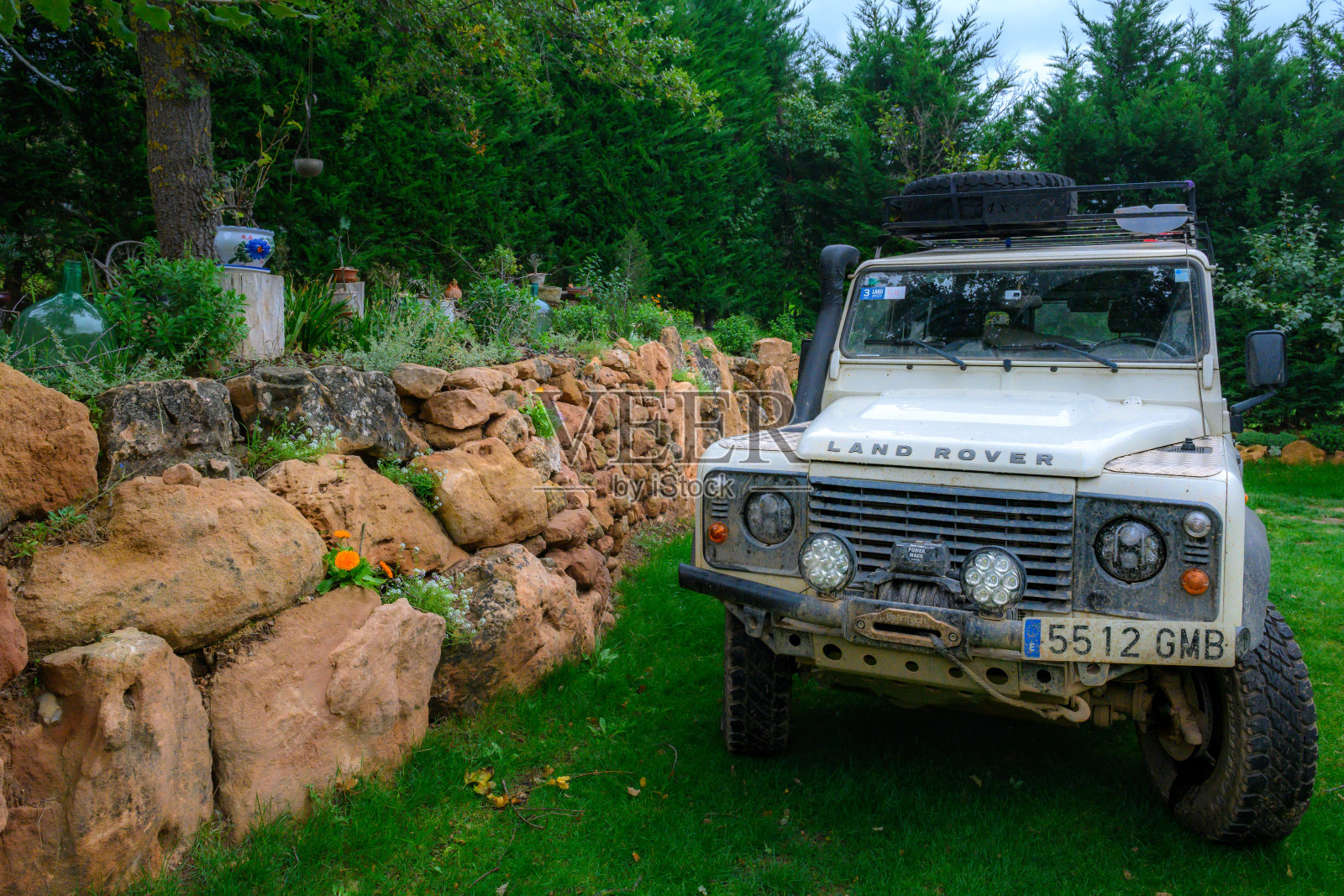 正面视角的白色坚固路虎卫士110 Puma，装备齐全，适合越野探险，停在生机勃勃的绿色草坪上。它身后是一面乡村风格的石墙和郁郁葱葱的植被，构成了一个风景如画的冒险背景。照片摄影图片