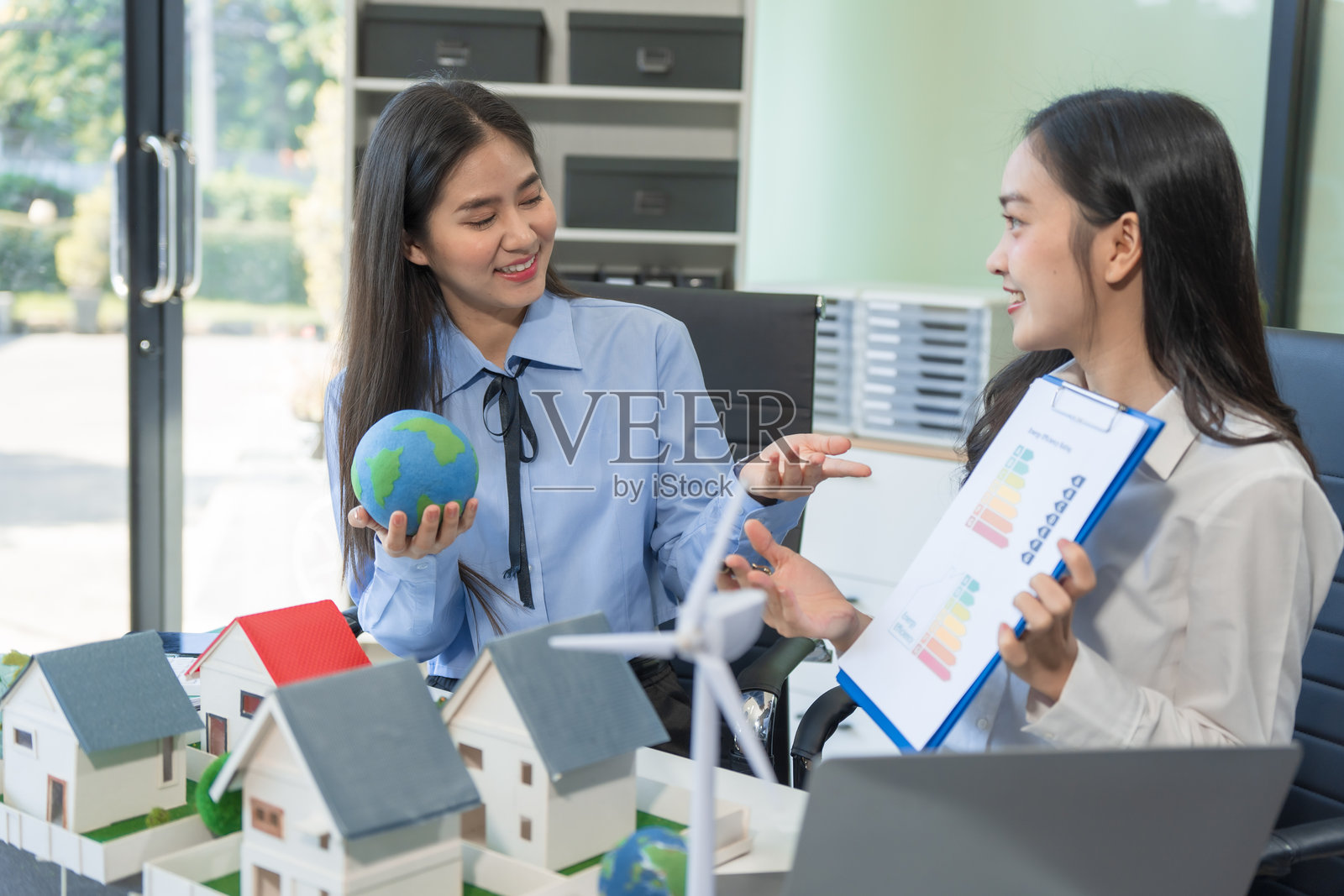 两位女商人在办公室的桌子旁坐着，讨论一个包含风车、绿色树木、可持续生活、可再生能源、循环经济和环保城市的模范村庄，为世界的未来而构思。照片摄影图片