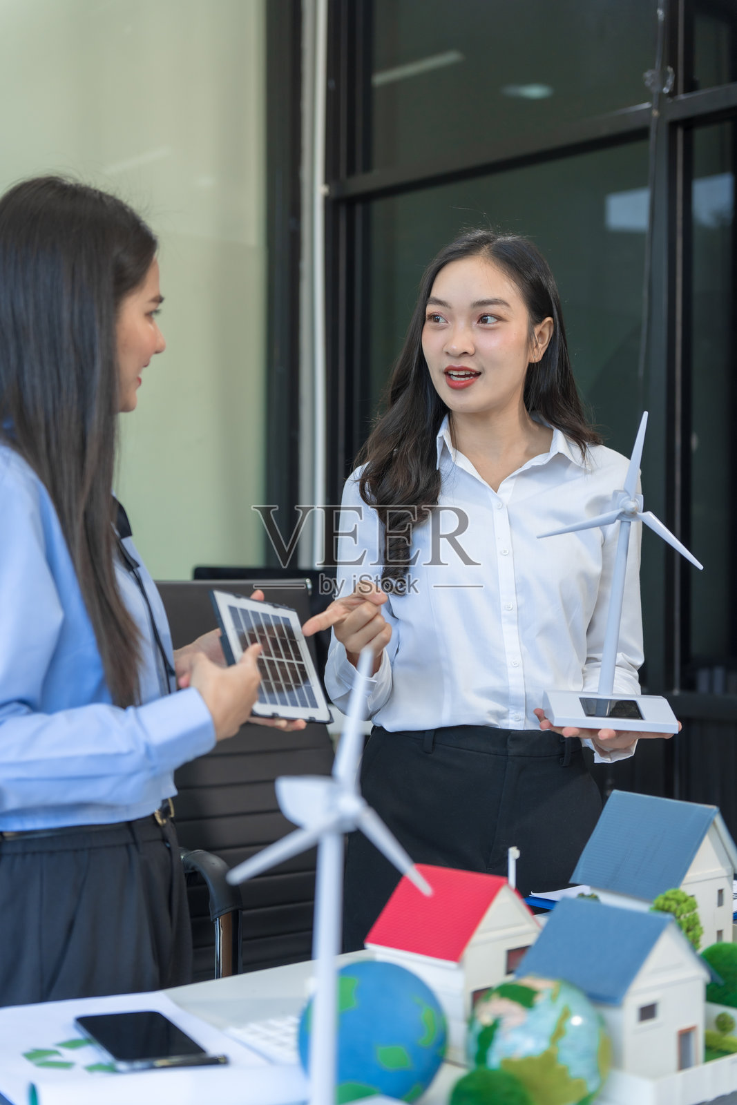 两位女商人在办公室的桌子旁坐着，讨论一个包含风车、绿色树木、可持续生活、可再生能源、循环经济和环保城市的模范村庄，为世界的未来而构思。照片摄影图片