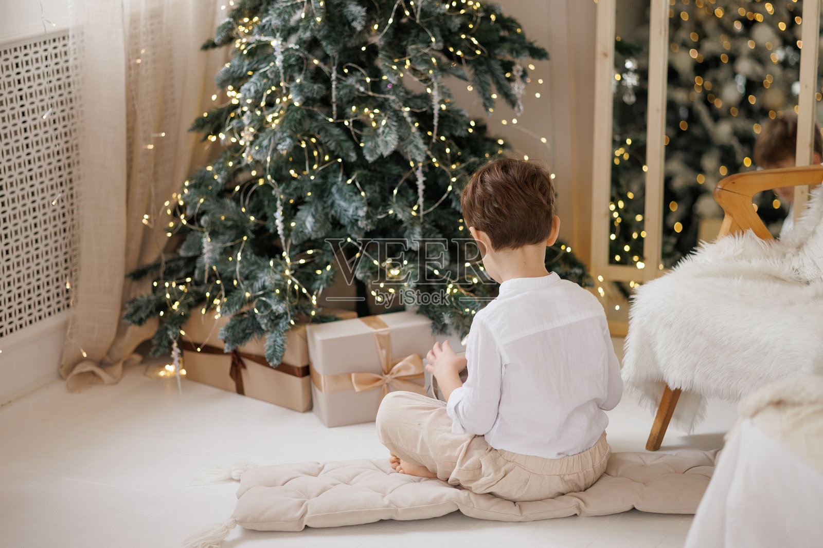 一个小男孩坐在床上，身穿白色衬衫，白色的毯子下，正在拆礼物。照片摄影图片