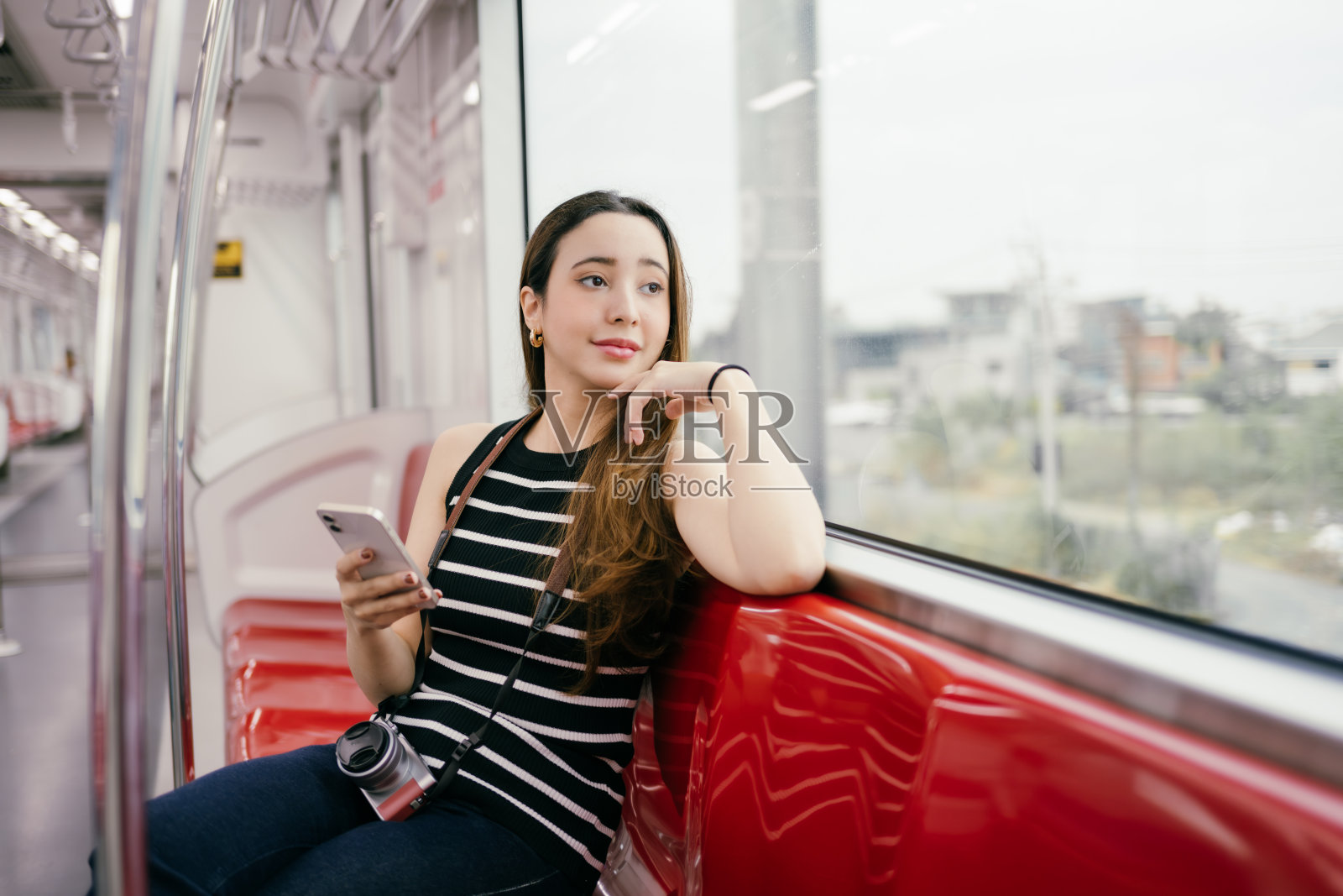 年轻女性游客舒适地坐在地铁上，手握智能手机，凝视窗外，享受日常通勤中的城市风景。照片摄影图片