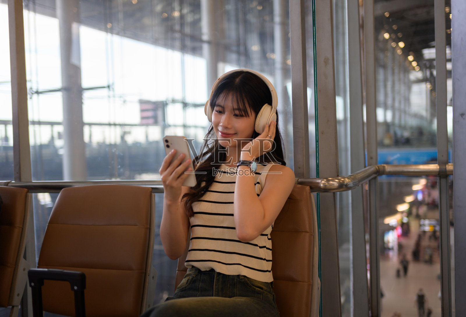 年轻的亚洲女性坐在机场休息室，戴着无线耳机，正在用智能手机听音乐或播客，等待她的航班。照片摄影图片