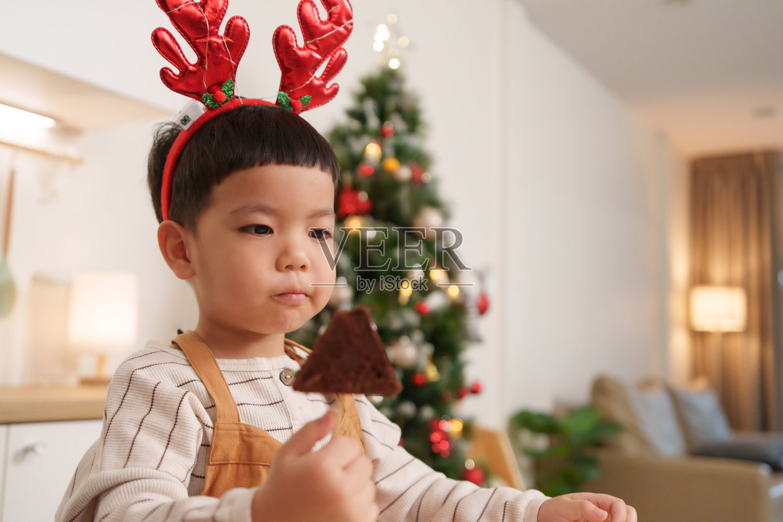 亚洲小孩在装饰圣诞饼干的同时吃饼干，客厅里装饰着圣诞树，洋溢着节日的欢乐气氛。照片摄影图片