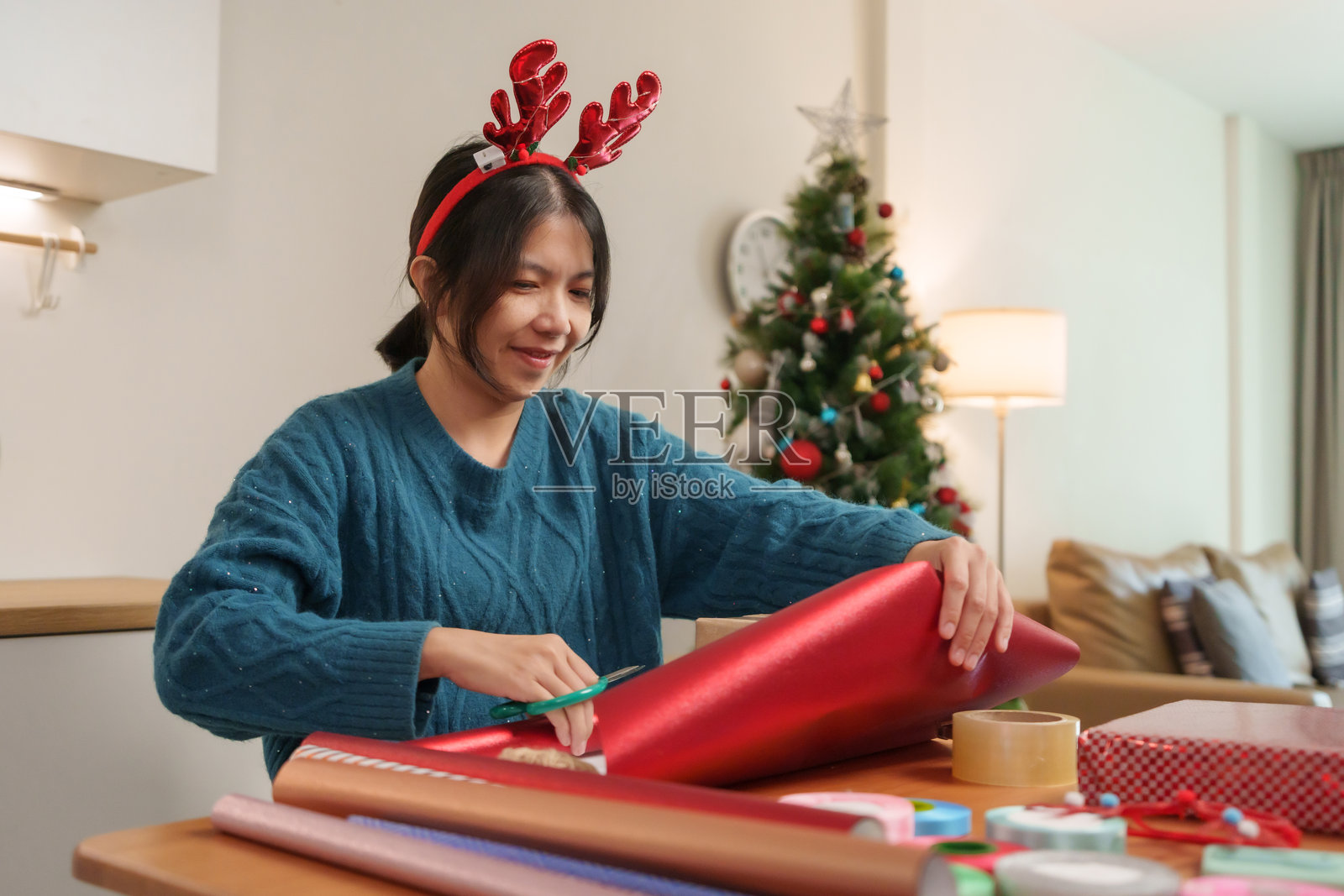 亚洲女性戴着驯鹿角，专注地包裹一个红色礼物，周围是节日装饰和一棵温馨的圣诞树。照片摄影图片