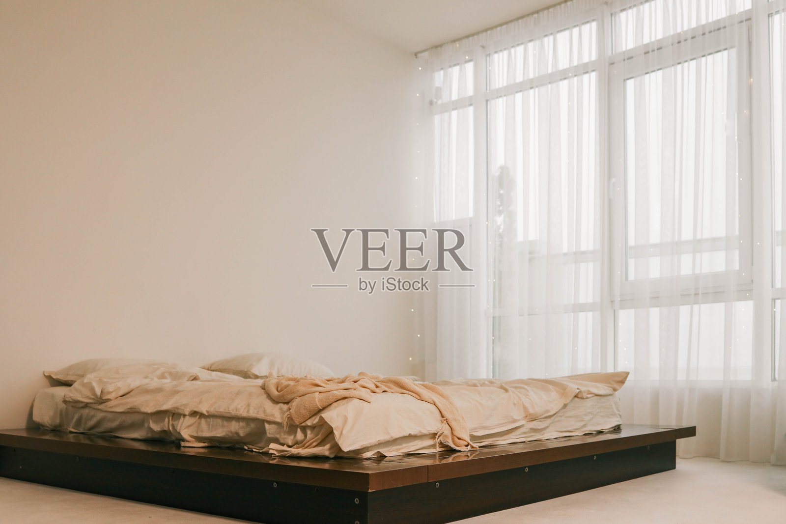 极简主义卧室，舒适的未整理床铺，柔和的米色调，大窗户上悬挂着轻薄窗帘，透入自然光线。宁静而安详的室内环境。照片摄影图片