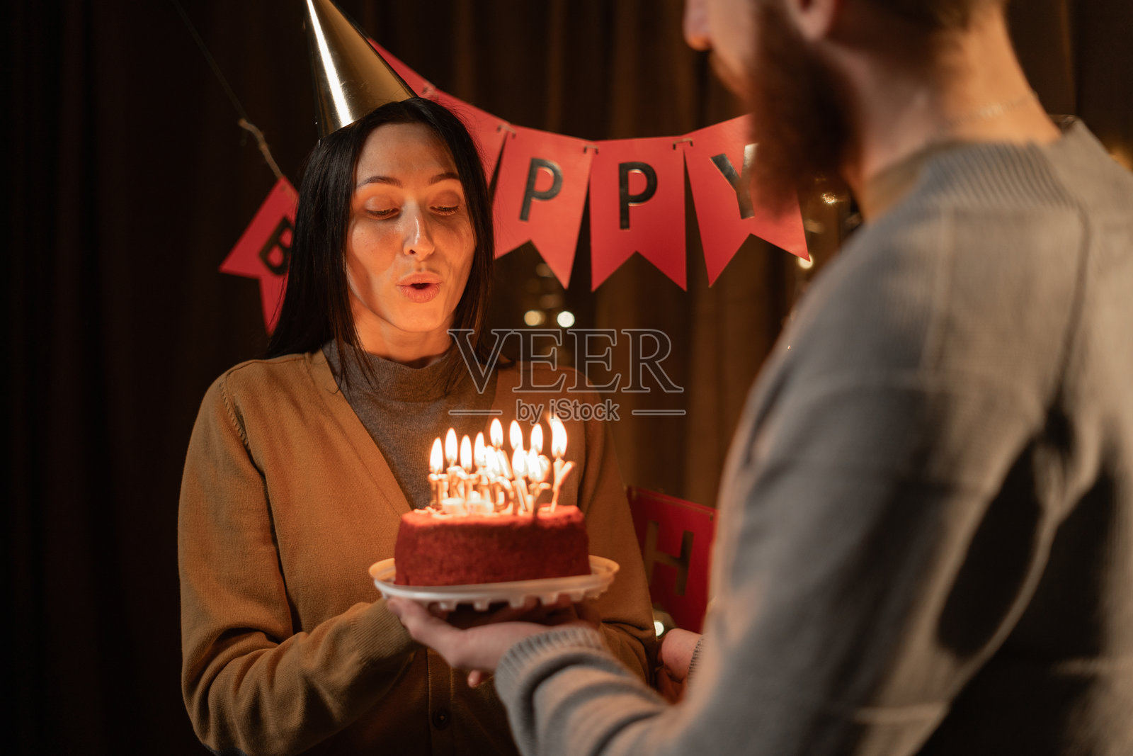 丈夫在晚上客厅里给妻子送上点燃蜡烛的蛋糕，妻子许下心愿并吹灭蜡烛。照片摄影图片