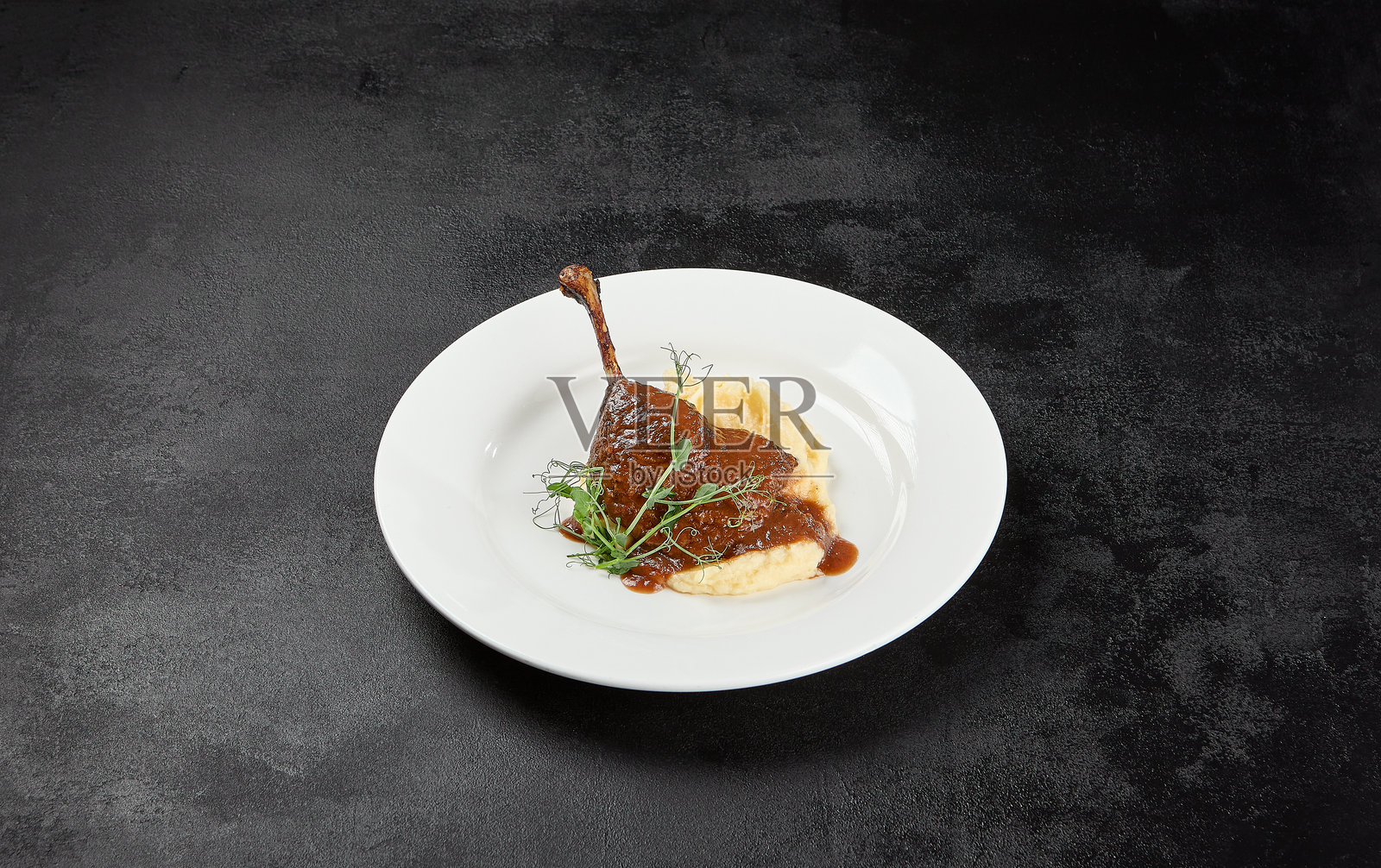 经典法式菜肴 - 黑色背景下的烤鸭腿配酱汁和装饰。深色混凝土桌上白色盘子里的熟鸭腿配土豆泥。极简风格的鸭肉油封。照片摄影图片