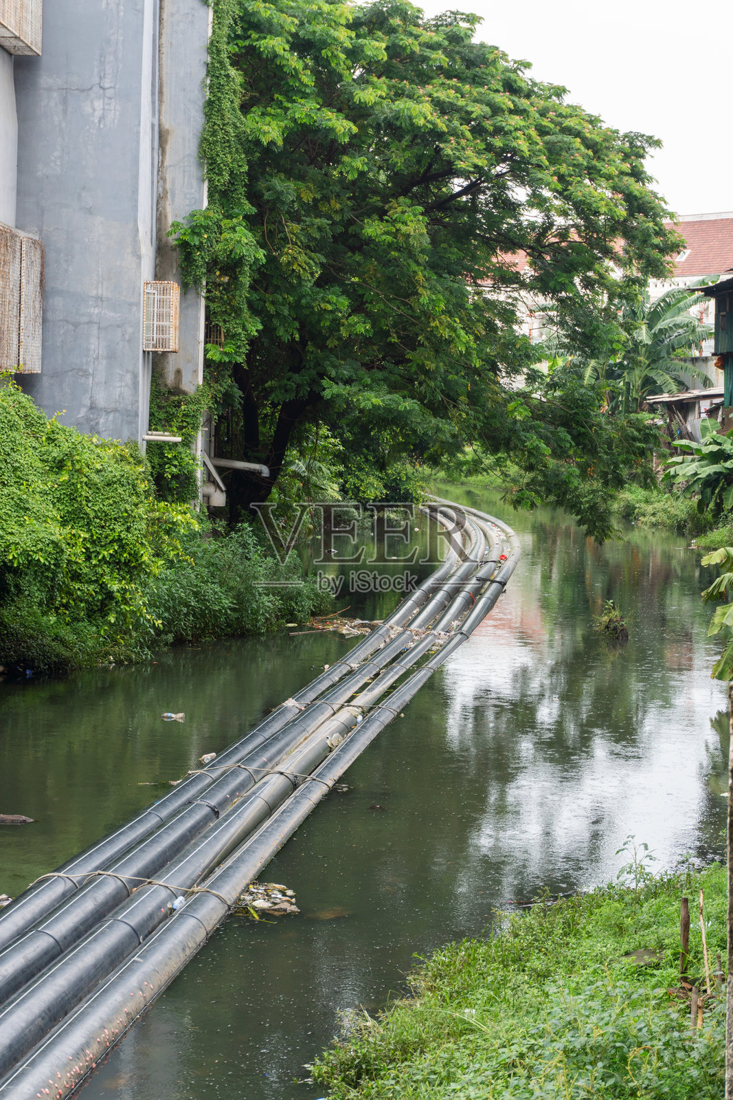 一条有管道的运河穿过城市建筑和郁郁葱葱的树木之间。照片摄影图片