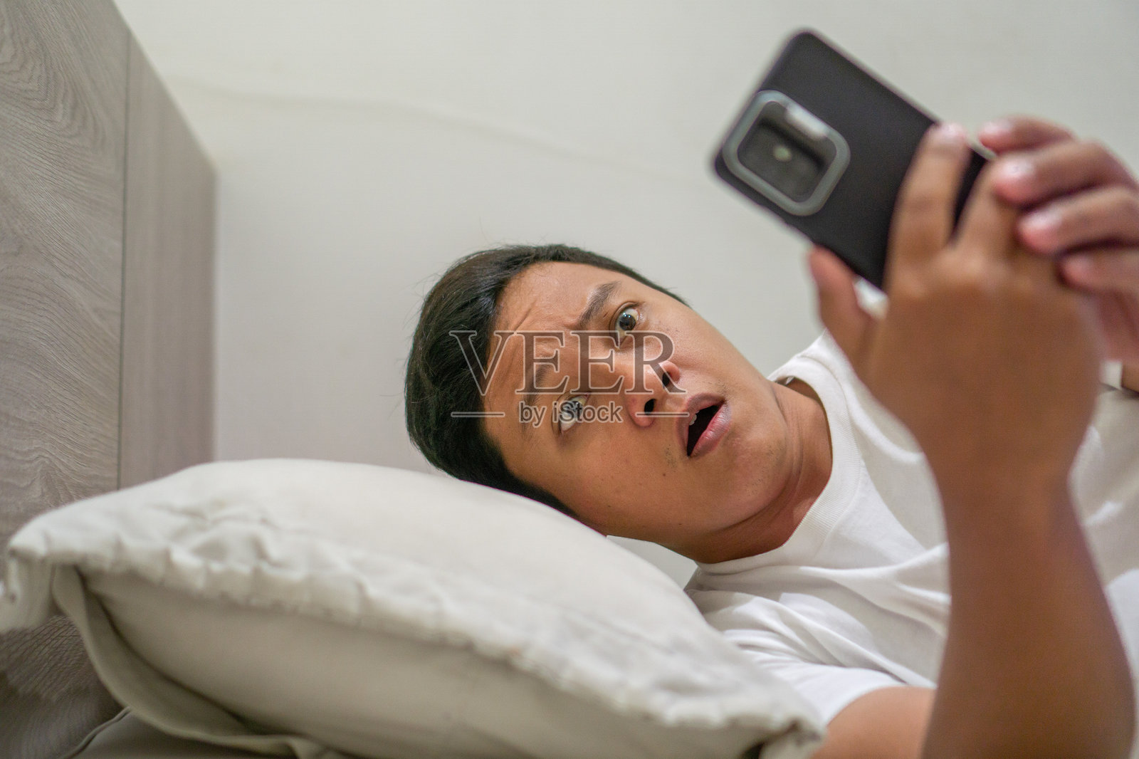 男子在床上夜晚拿着智能手机，露出惊讶的表情。照片摄影图片