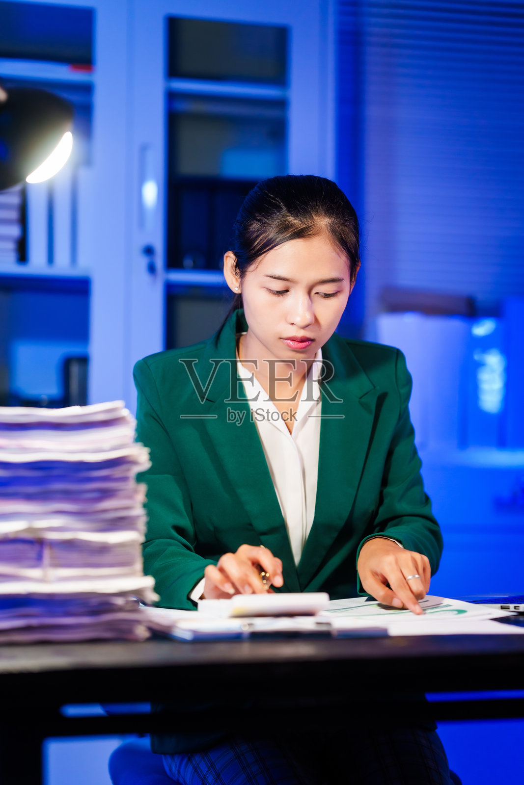 一位亚洲女商人在夜晚加班，坐在办公桌前，通过电脑或笔记本电脑查阅大量文件。面对办公室问题，她一边处理繁重的工作，一边在线聊天。照片摄影图片