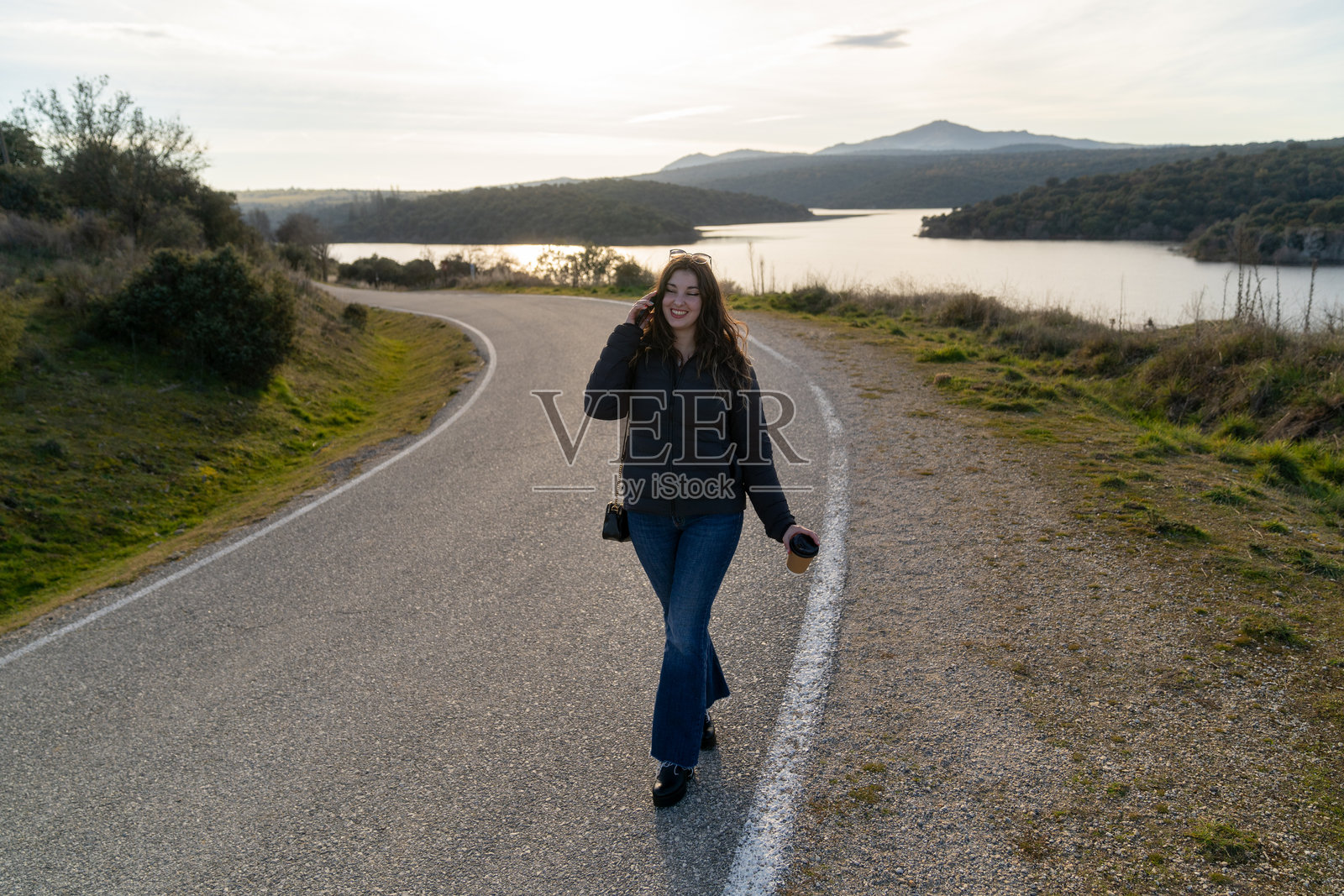 年轻女性在湖边的乡间小路上走，手里拿着咖啡和手机照片摄影图片