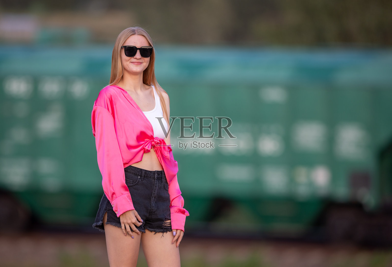 年轻女性肖像，戴着太阳镜，穿着红色衬衫，长长的金发，背景是货运列车车厢。照片摄影图片