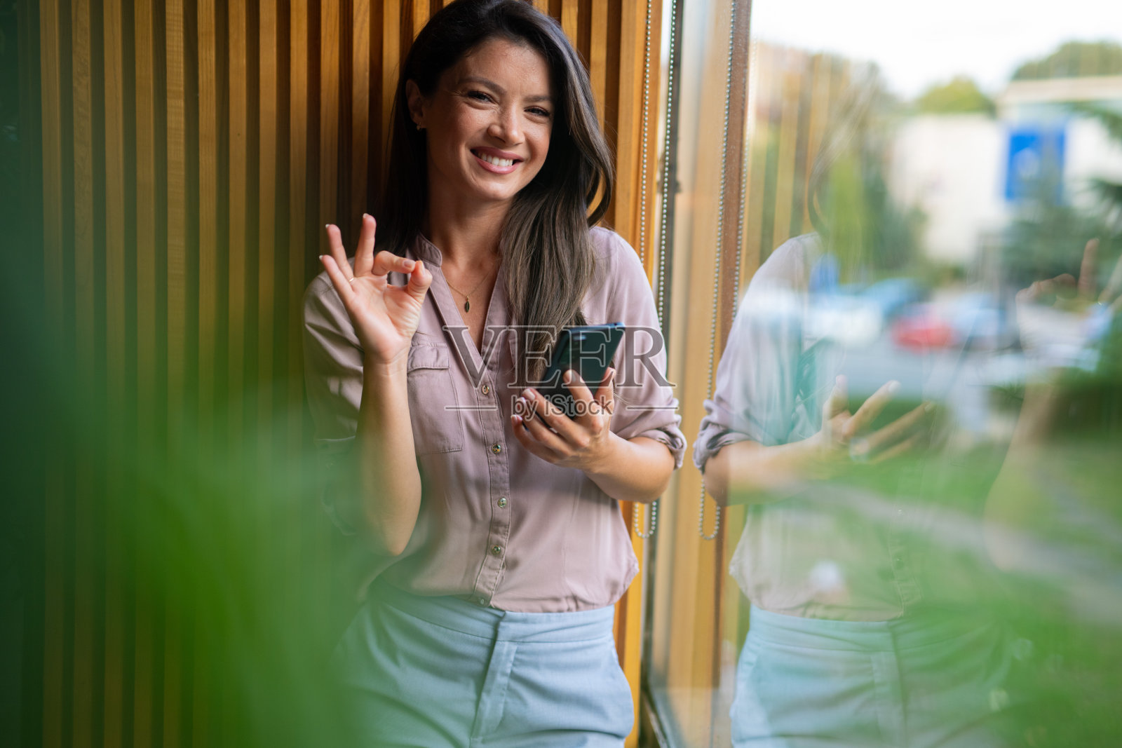 商务女性做出“好”的手势并拿着手机。女性站在窗边的肖像。照片摄影图片
