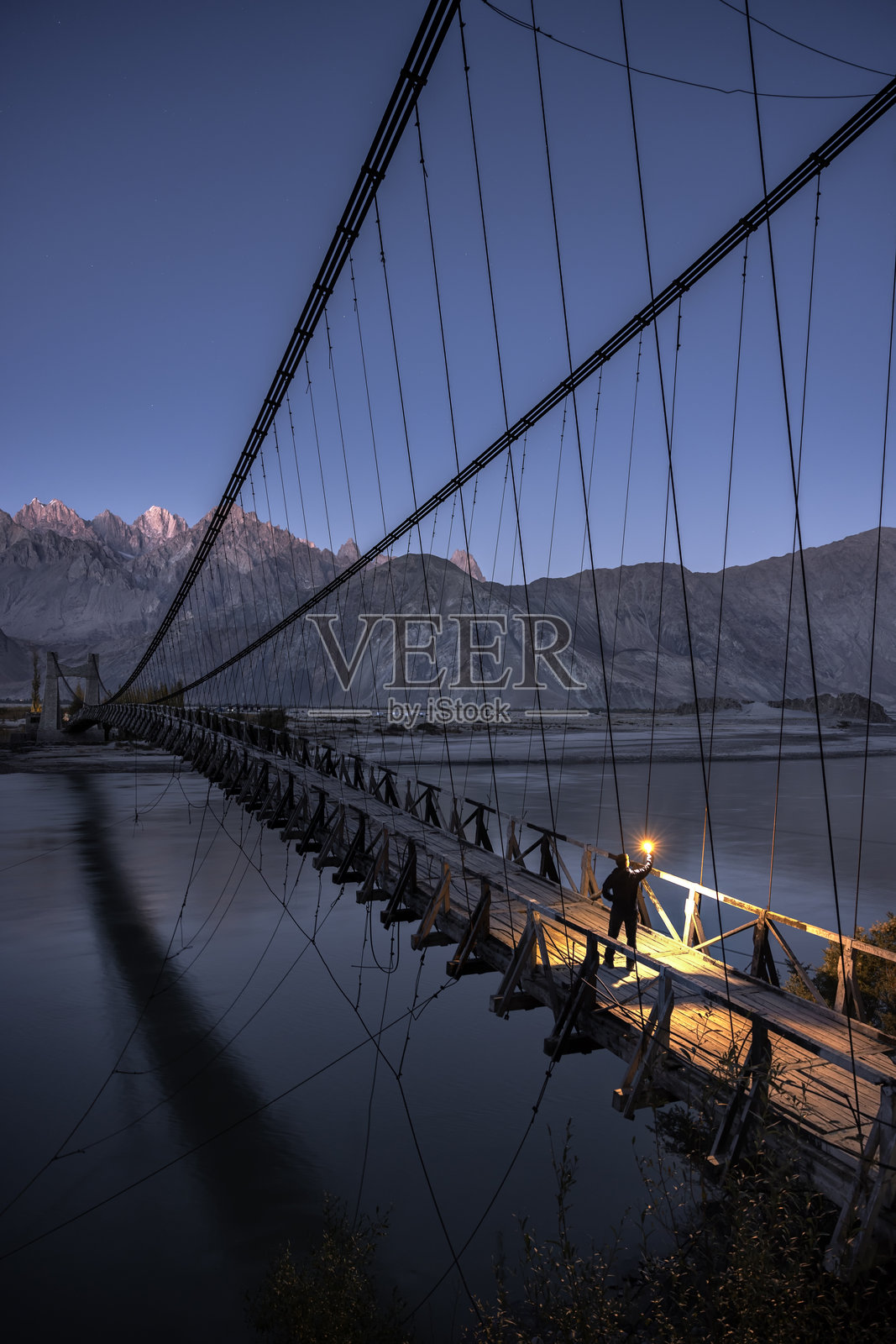 一名男子在日落时分手持灯笼,走在桥上穿越巴基斯坦斯卡杜的印度河。照片摄影图片