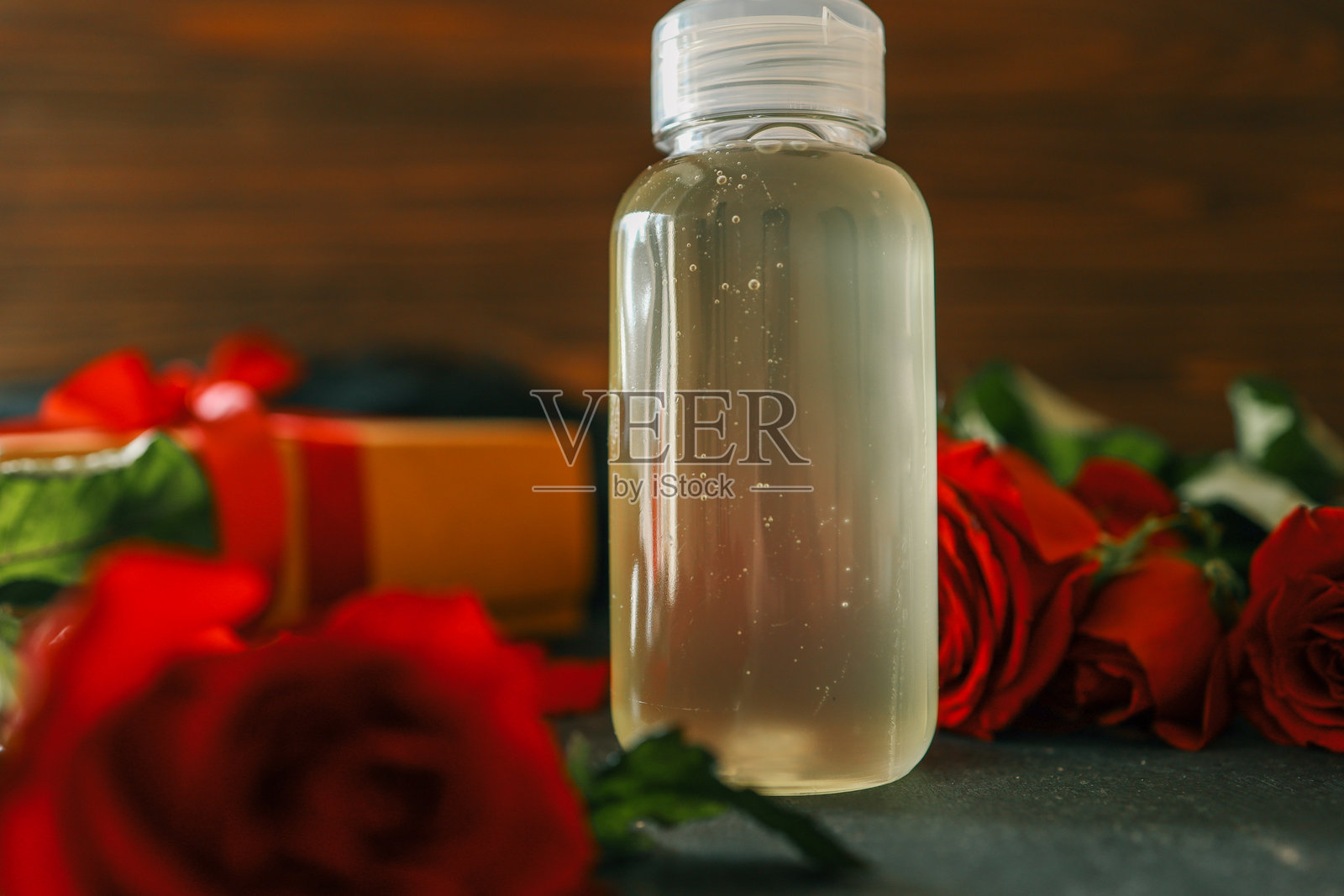 透明瓶中装有清澈液体，放置在黑色表面上，周围环绕着红玫瑰和一个系有红色丝带的礼品盒，背景为木质纹理，营造出温暖自然的美感。照片摄影图片