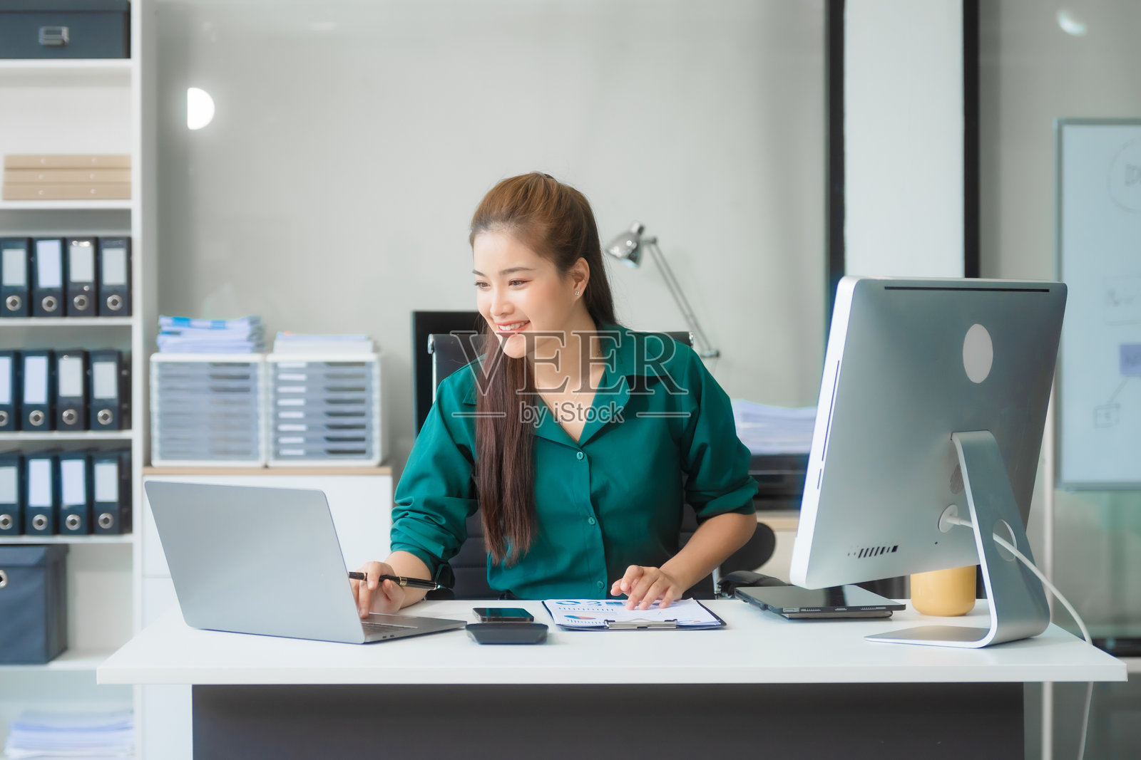 身穿正式商务服装的迷人亚洲女商人在办公桌前使用笔记本电脑和手机，专注于公司法、合同、合规以及律师事务所的营销策略。照片摄影图片