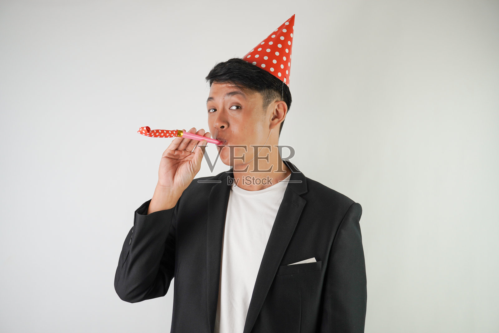 兴奋的年轻亚洲男子戴着红色派对帽，穿着白衬衫和黑色西装，正在吹派对喇叭，背景为白色。适合圣诞节和新年派对的概念。照片摄影图片