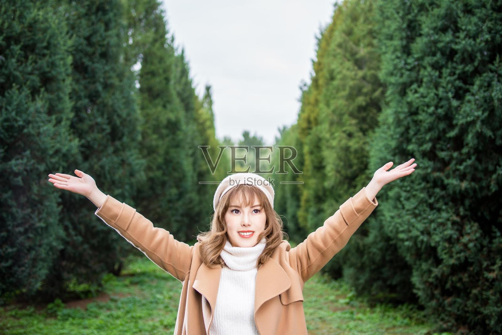 一位年轻女性穿着棕色外套和白色高领毛衣，双臂伸展站在森林中。照片摄影图片