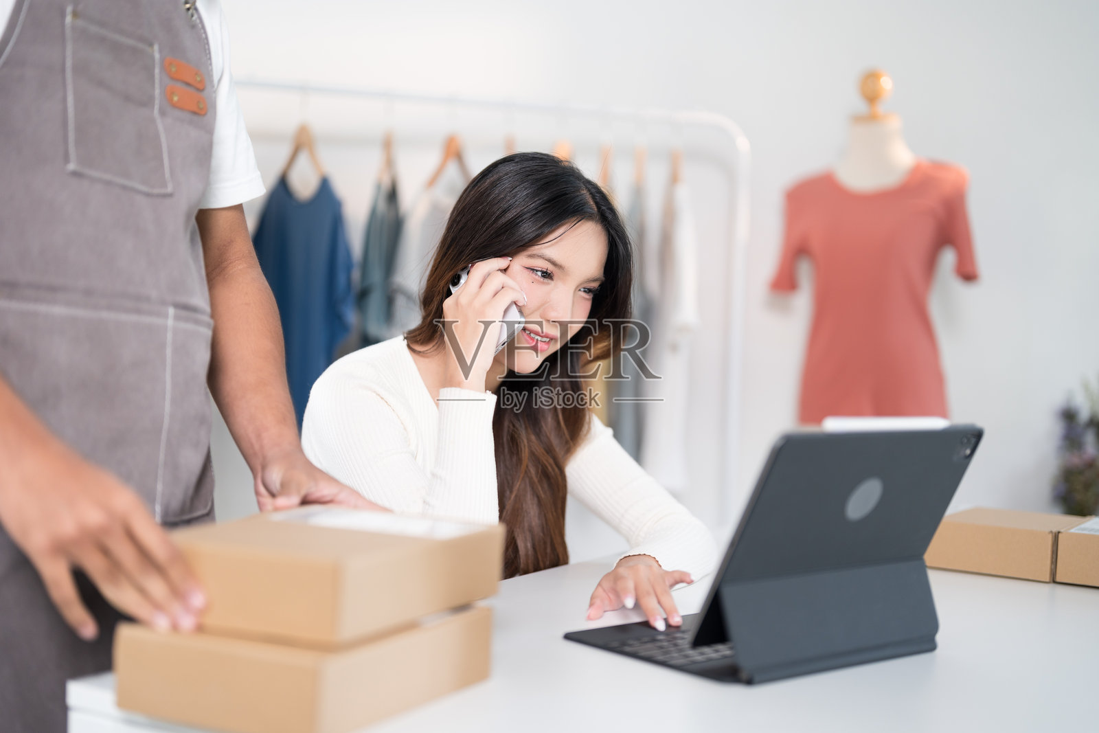 一位女性在工作空间中一边打电话，一边使用笔记本电脑，周围是箱子和衣架。照片摄影图片
