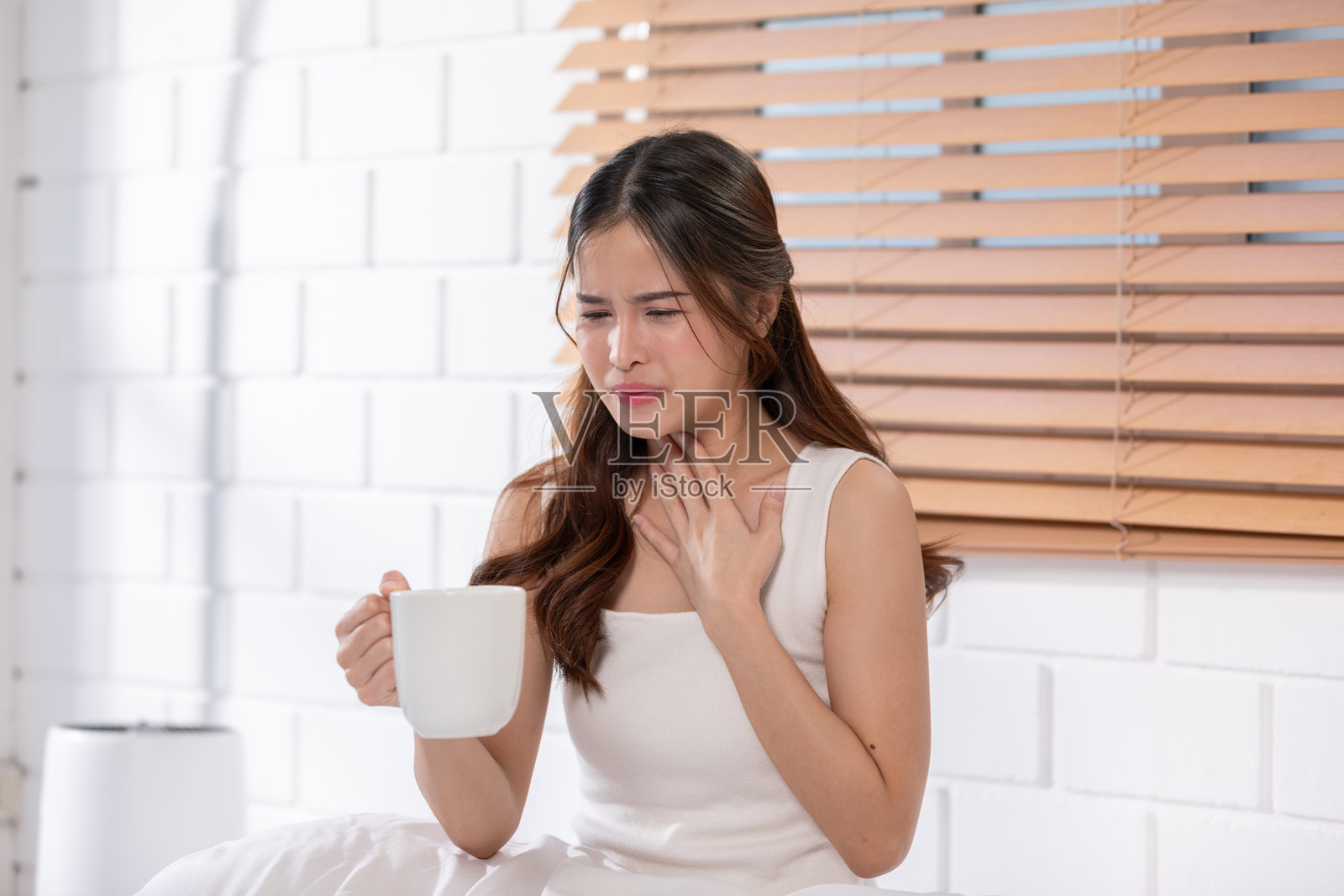 穿着白色背心的亚洲女性手持杯子，表现出不适并触摸喉咙，健康护理概念照片摄影图片