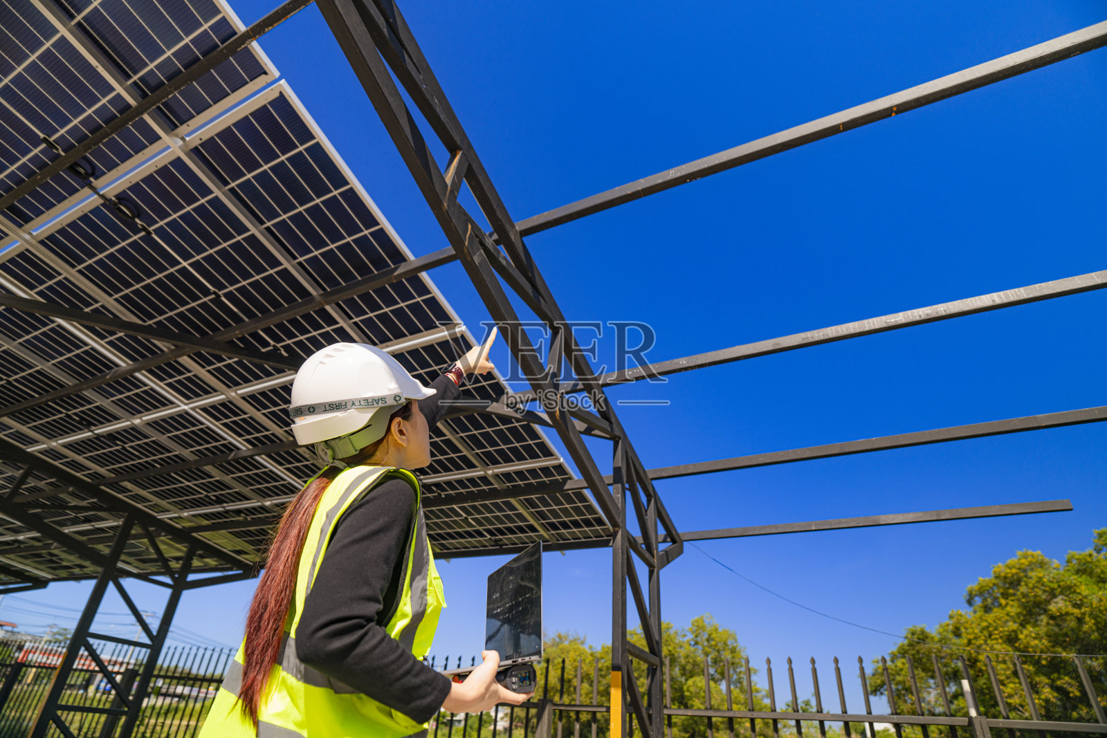 一位穿着安全背心和头盔的女性工程师正在检查一座正在建设中的太阳能车棚。结构的钢梁和部分安装的太阳能板在明亮的蓝天下清晰可见。照片摄影图片