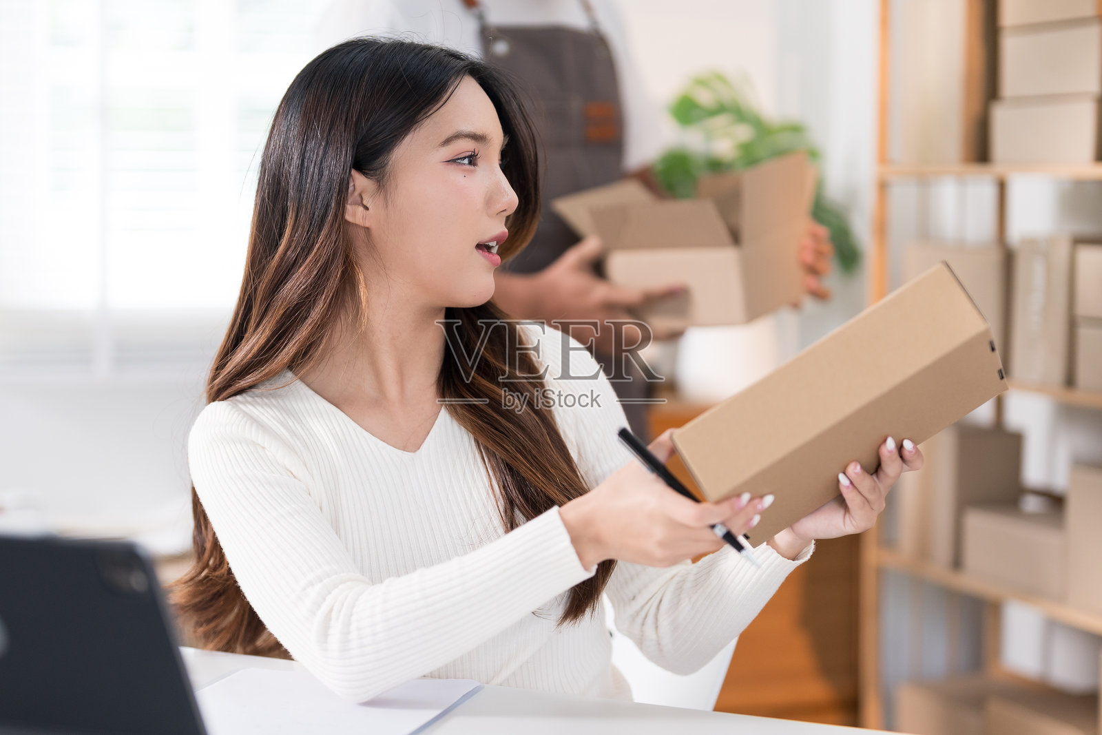 一位女性热情地进行直播，手中拿着一个盒子，展示她的产品，工作空间里有环形灯和笔记本电脑，四周摆满了货架和包装。照片摄影图片