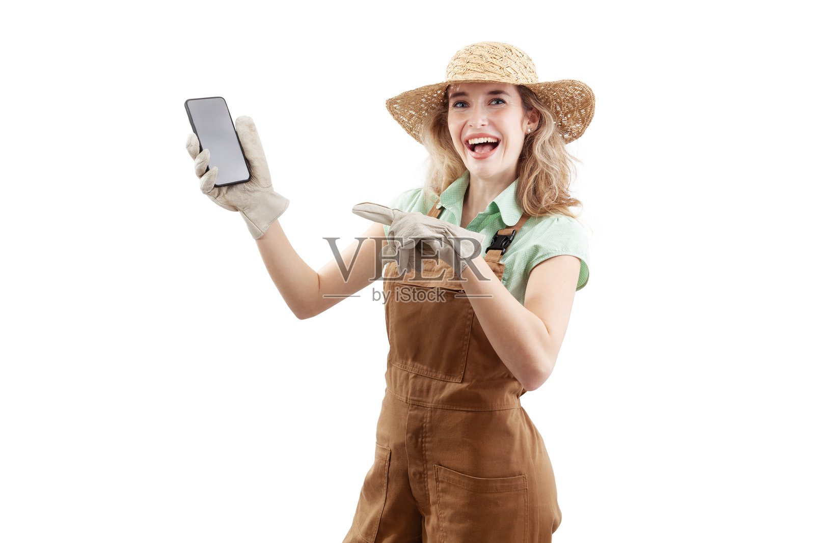 微笑的女性穿着围裙、帽子和园艺手套，背景为白色，指着一部智能手机。在线园艺购物，为春天的到来和园艺工作做准备。照片摄影图片