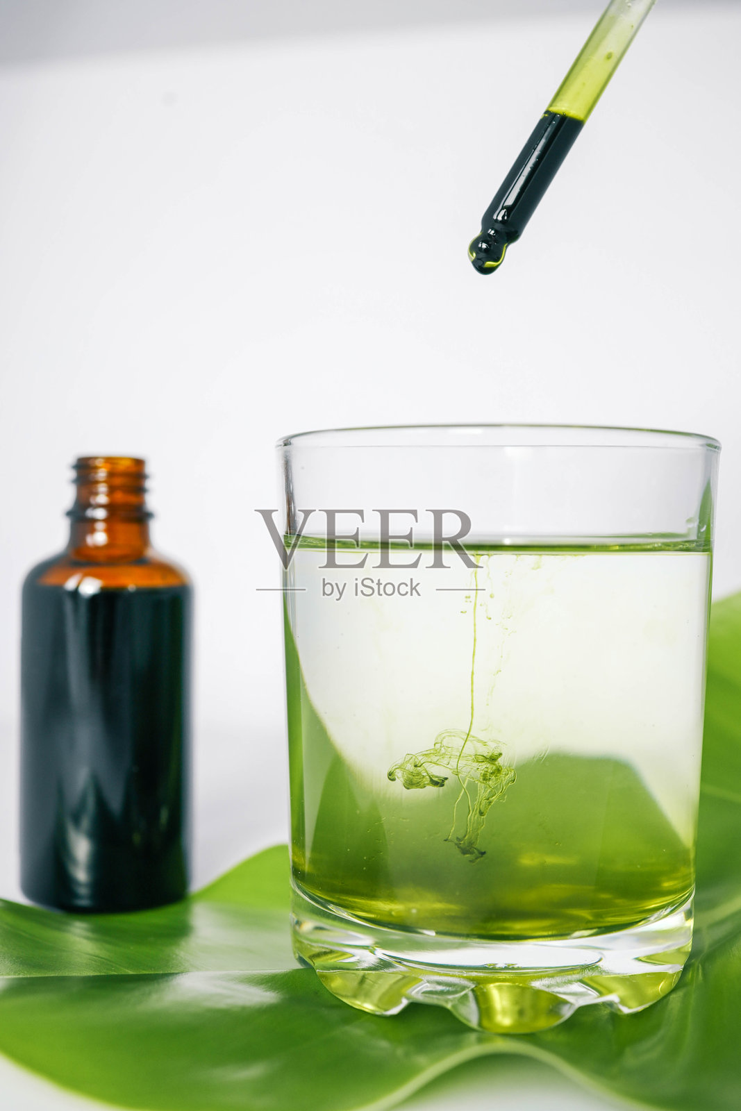 滴管将绿色液体滴入水杯中，水杯下方有一片叶子，背景中有一个棕色玻璃瓶。代表天然成分和有机产品。照片摄影图片