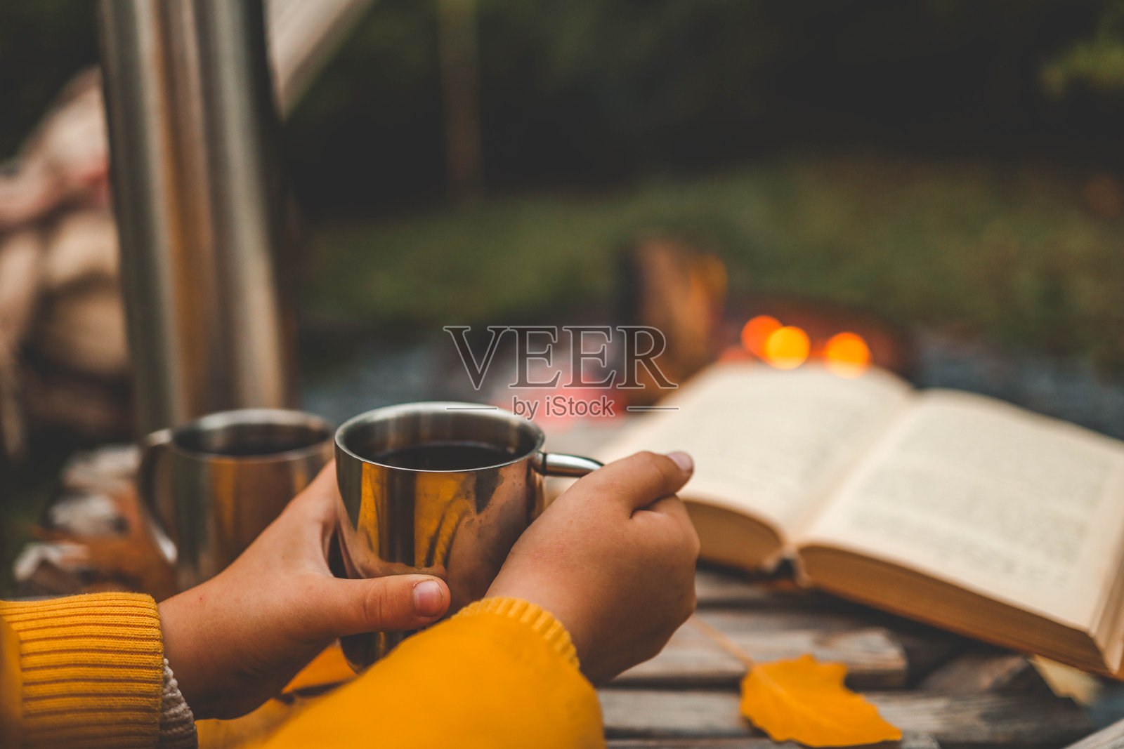一个舒适的户外场景，描绘一个人手握金属杯中的热饮，坐在篝火旁，旁边有一个保温瓶、一本书和秋天的落叶，散发着宁静的秋日氛围。照片摄影图片