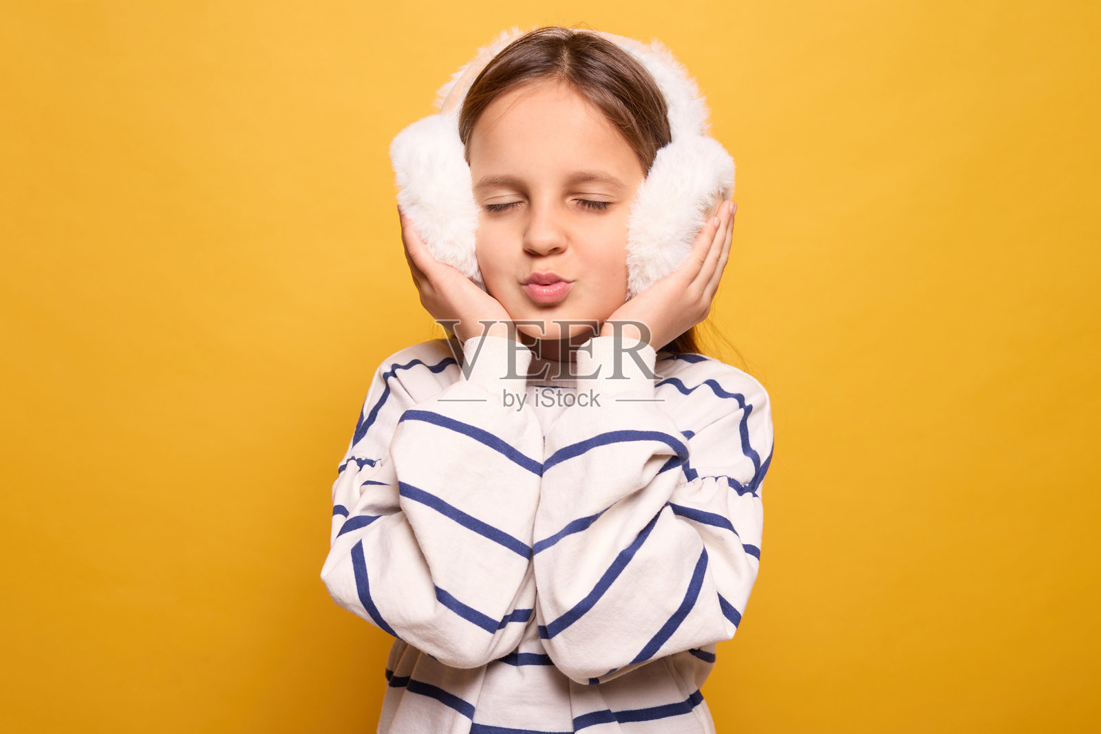 一个长发女孩在柔软的耳罩中顽皮地摆姿势，闭着眼睛，嘴唇微微嘟起，享受着温暖，背景是鲜艳的黄色。在这个生动的环境中，冬季时尚熠熠生辉。照片摄影图片