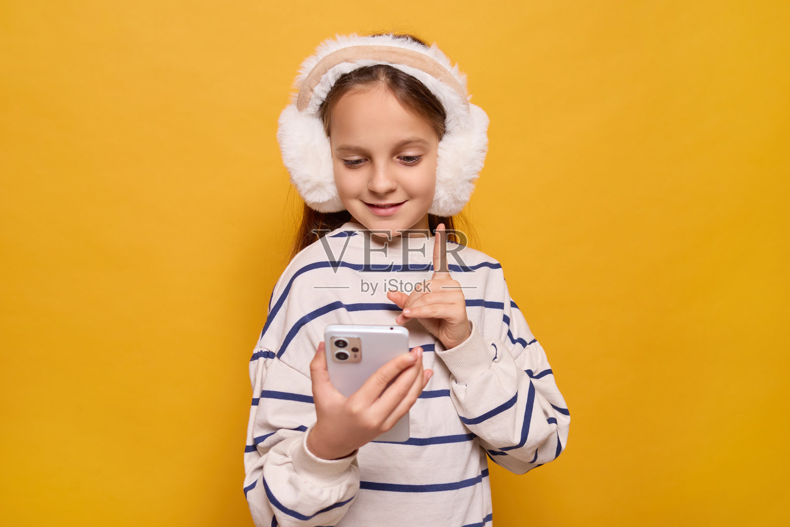 一个年轻女孩专注于她的智能手机，微笑着用手指做出手势，戴着舒适的耳罩，背景是充满活力的黄色。照片摄影图片