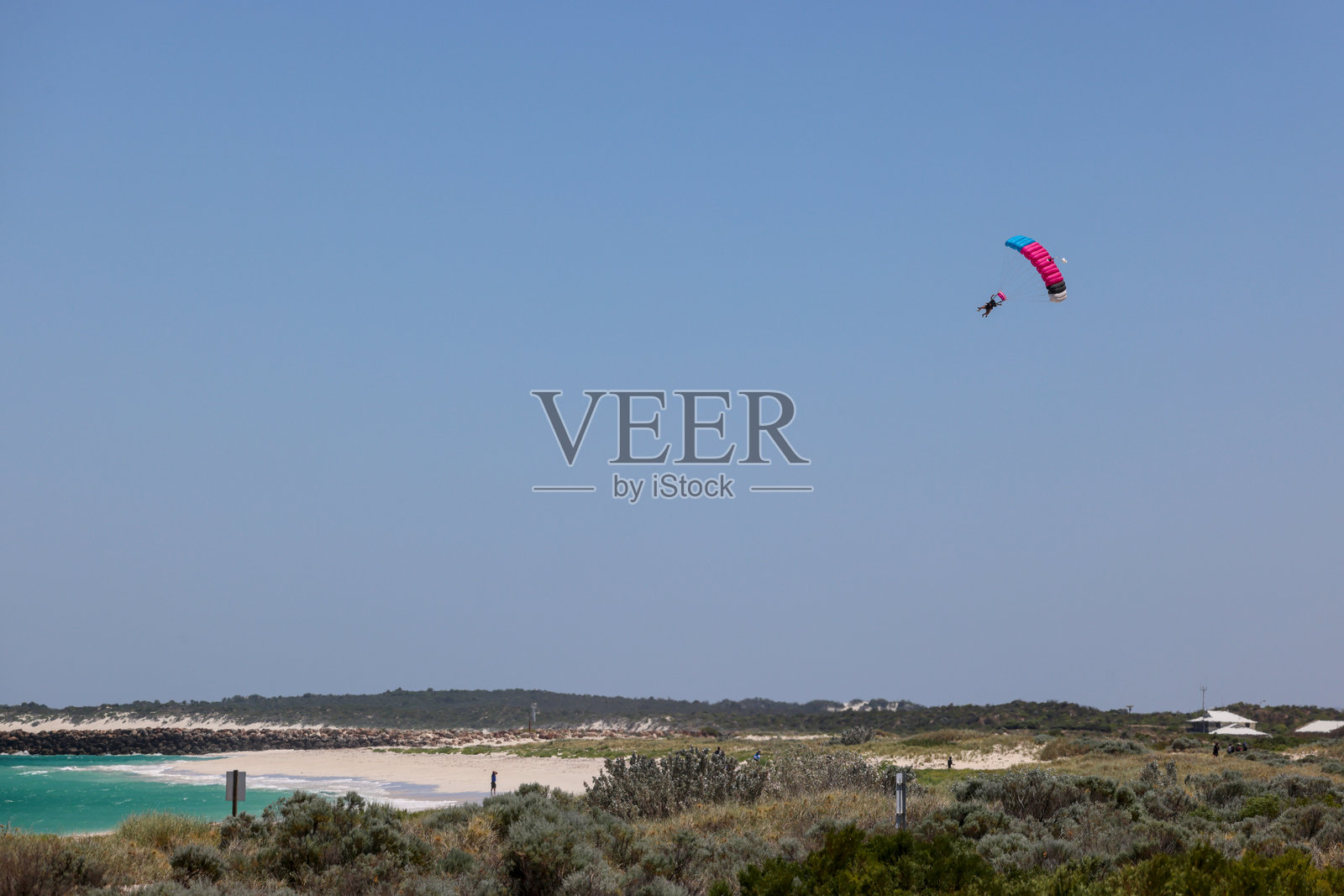 在晴朗的蓝天上进行双人跳伞，降落在白色沙滩上，海水呈现出碧绿色。照片摄影图片