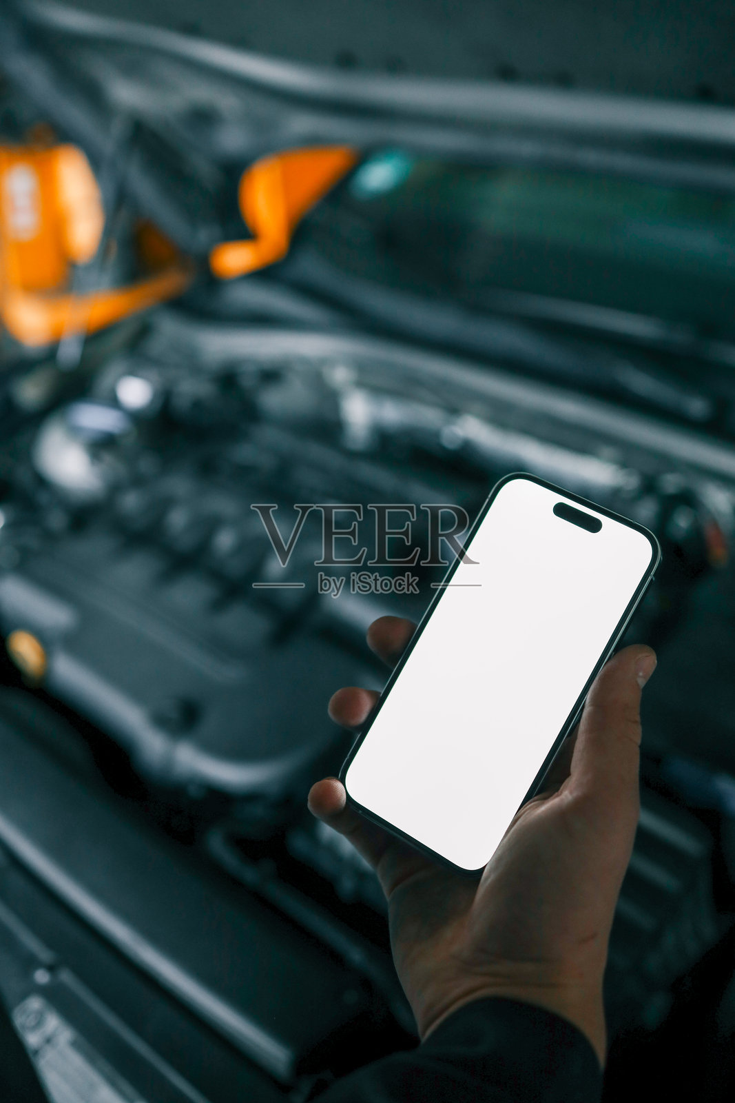 机械师手持一部白屏智能手机，站在打开的汽车引擎前进行诊断或维护。室内车间环境。照片摄影图片